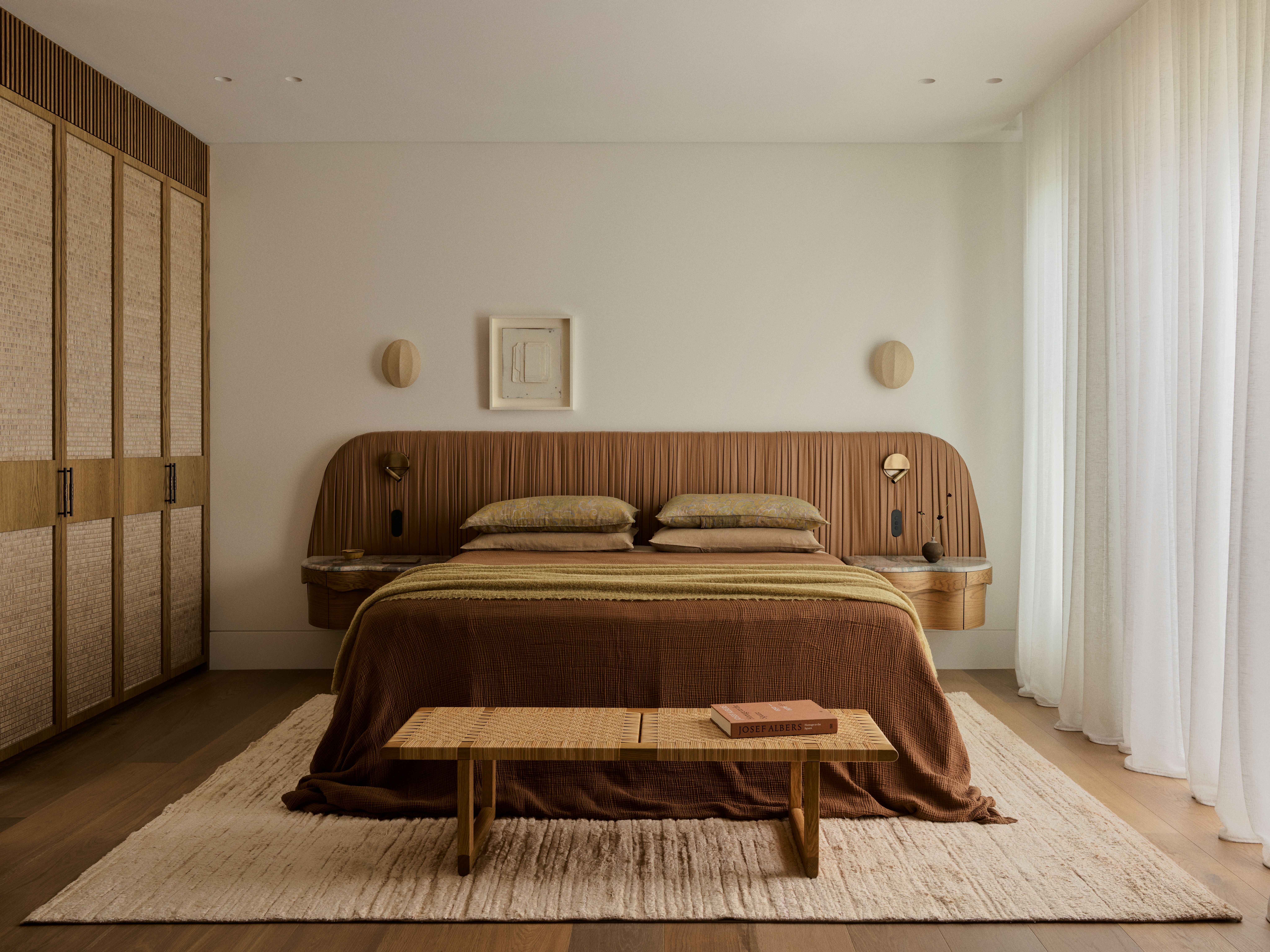 Dancing On The Ceiling Bedroom with a curved, ribbed leather headboard incorporating built-in nightstands, layered brown and olive bedding, a woven bench at the foot of the bed, and rattan wardrobe doors beside sheer curtains