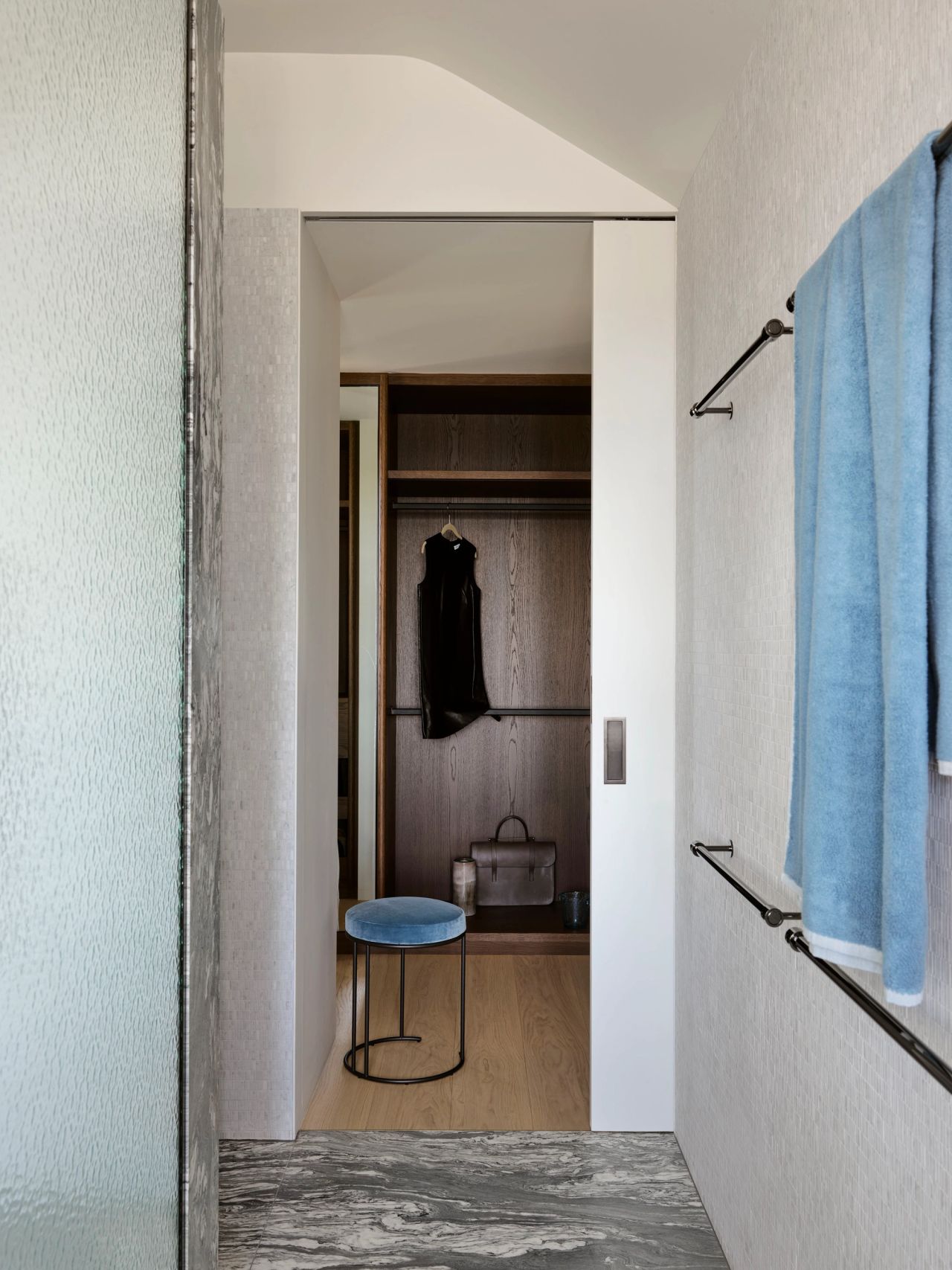 Contemporary master ensuite by Decus featuring textured white tiles, grey veined stone flooring, blue towels and a dark timber walk-in wardrobe with custom joinery.