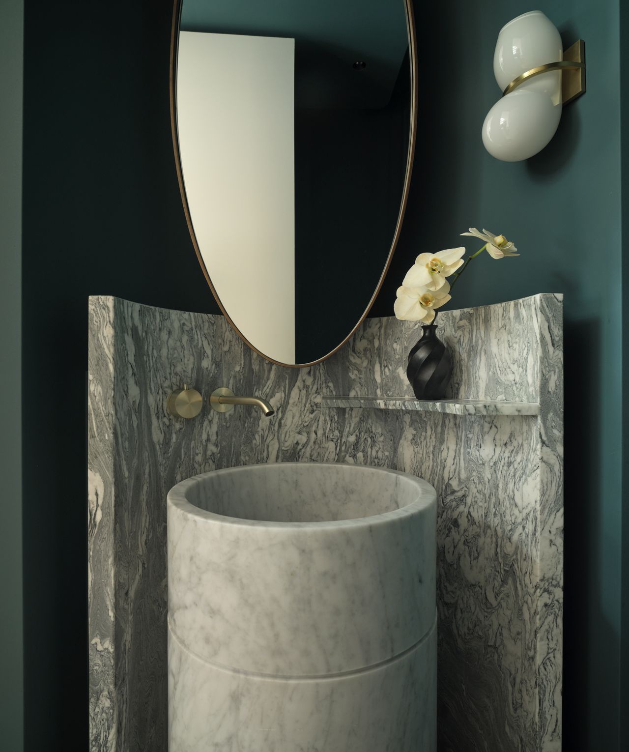 Monsieur G Powder Room Sculptural round marble pedestal basin set against a book-matched grey marble splashback, with an oval brass-framed mirror above and a brass double-globe wall sconce to the side, styled with a black ceramic bud vase and white orchid stem