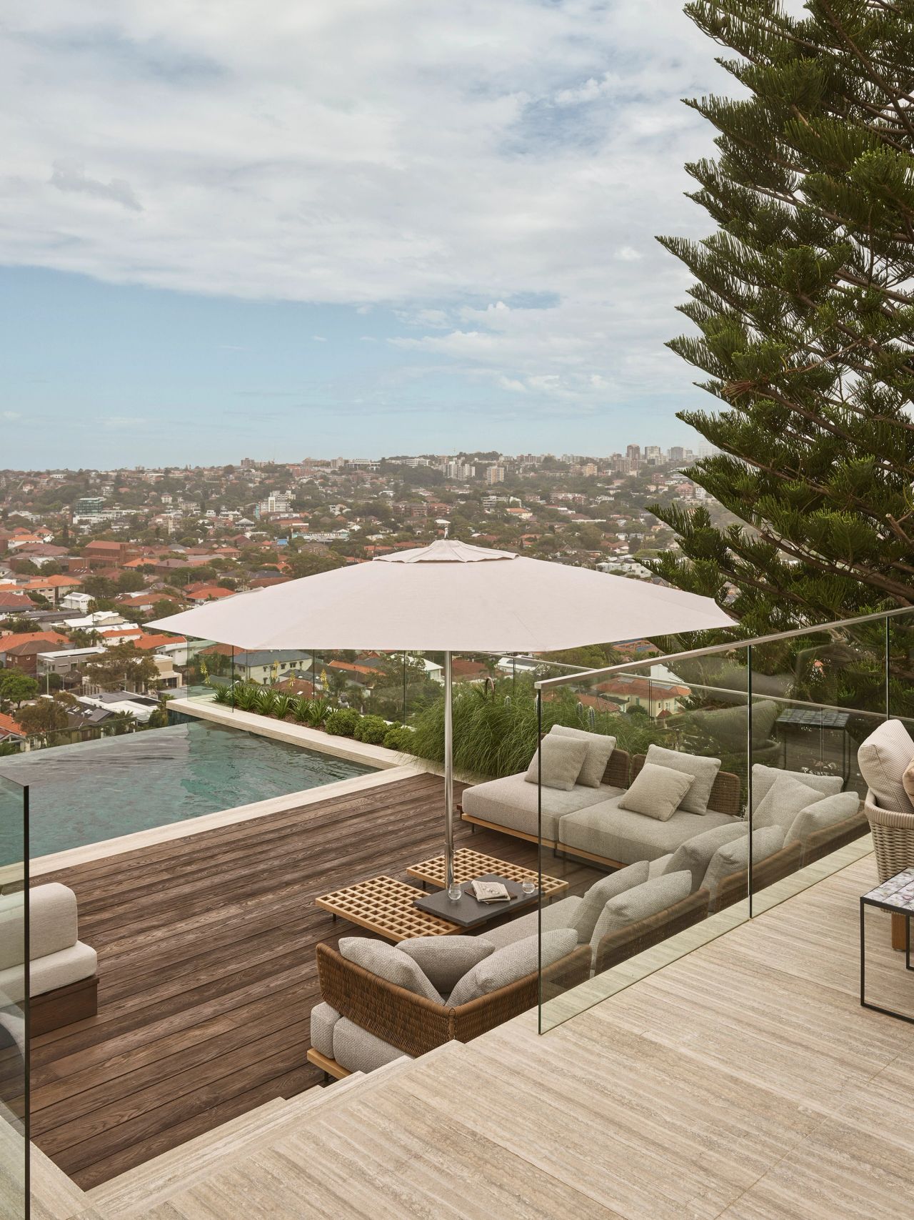 Orla outdoor timber terrace and pool view over the rolling hills of Sydney's eastern suburbs. Stone coloured ourdoor sofas and a cream coloured umbrella make a cozy outdoor lounge space - travertine flooring leading up to the timber decking a level below with the pool