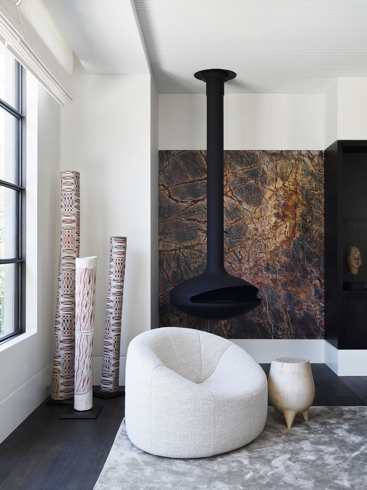 Contemporary living room with a suspended black fireplace, richly veined stone surround, sculptural white armchair and tall patterned artworks beside steel-framed windows.