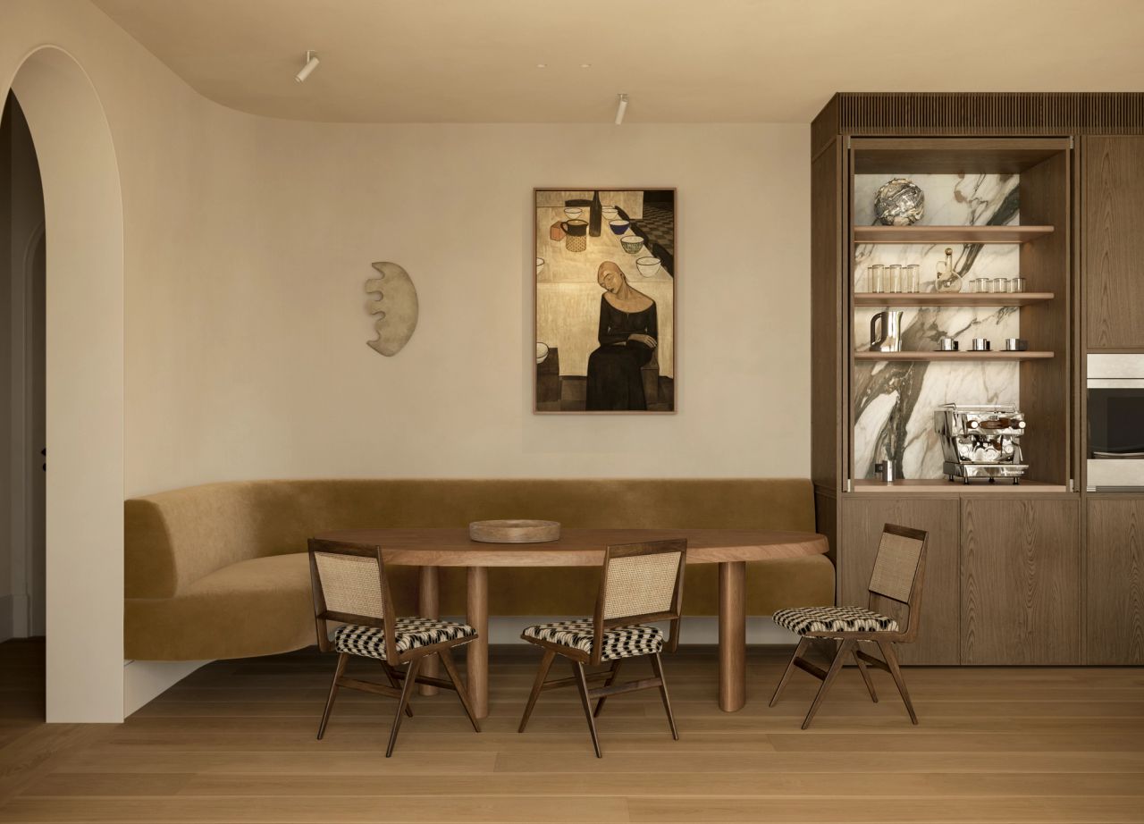 Lady M Dining nook with a built-in ochre banquette, oval timber table, and cane-back chairs upholstered in a graphic black-and-white weave, set against a marble-lined display cabinet and a figurative painting on the wall