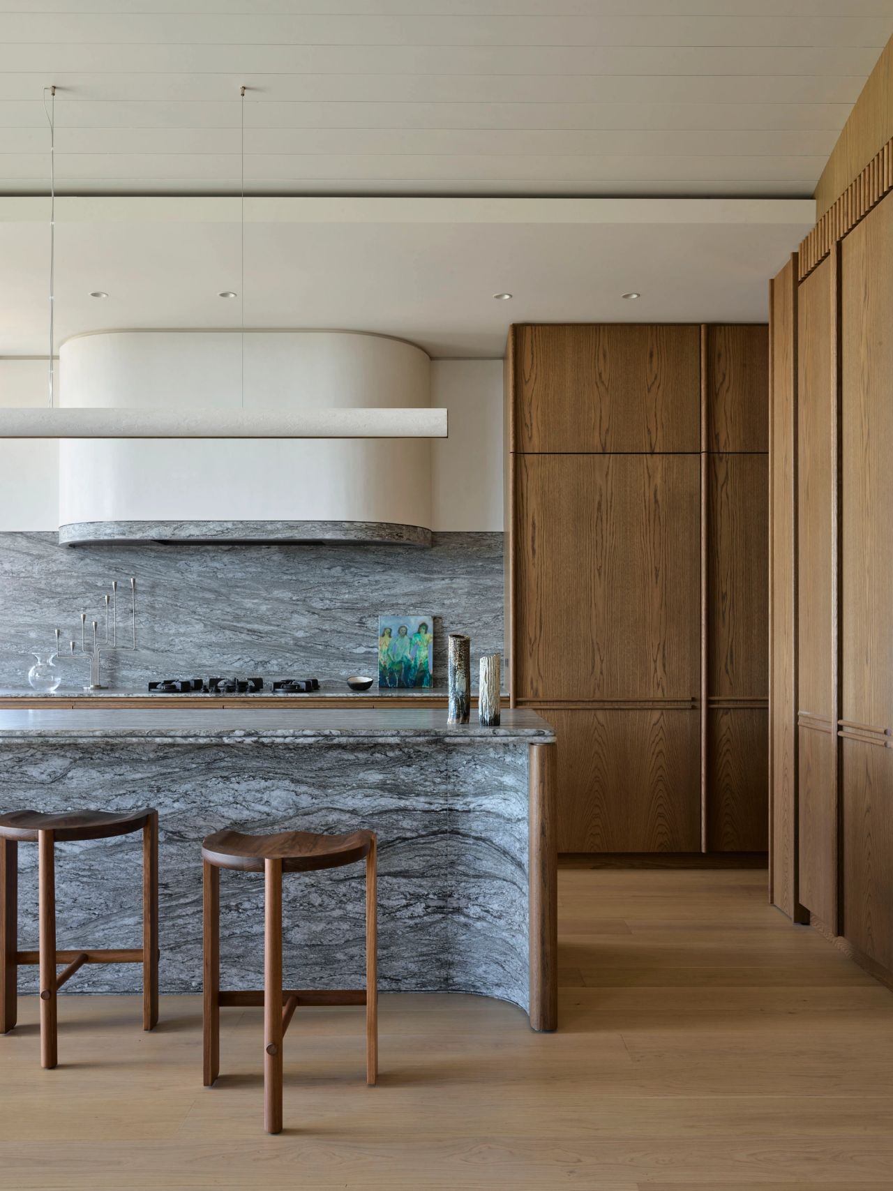 Ultramarine Kitchen with a curved grey marble island and matching splashback, a sculptural white pendant range hood, dark cooktop, wood bar stools, and floor-to-ceiling walnut cabinetry