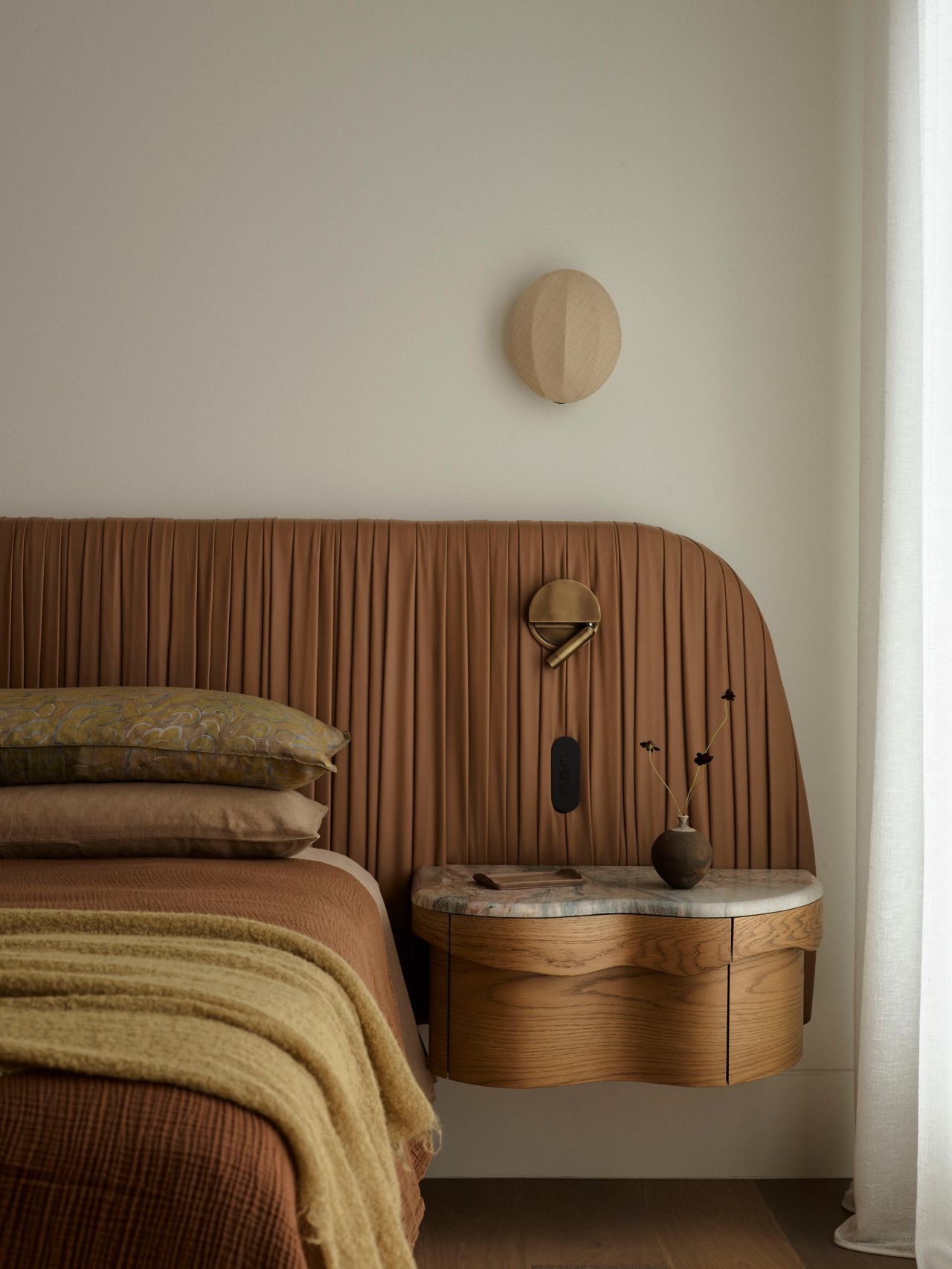 Dancing on the Ceiling Primary Bedroom with a curved, ribbed leather headboard incorporating built-in nightstands, layered brown and olive bedding, a woven bench at the foot of the bed, and rattan wardrobe doors beside sheer curtains