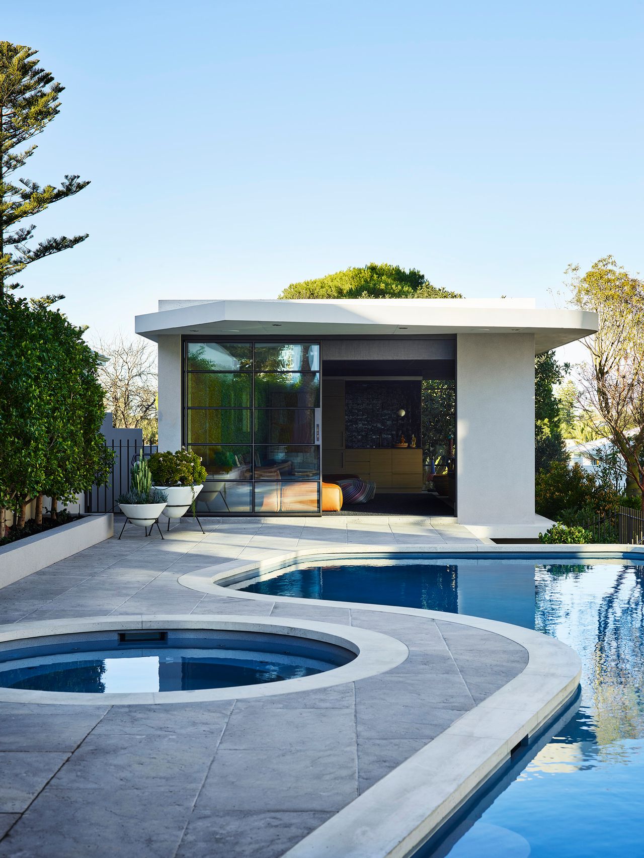 Curved outdoor swimming pool and circular spa beside a contemporary pool pavilion, surrounded by mature greenery and native coastal planting.