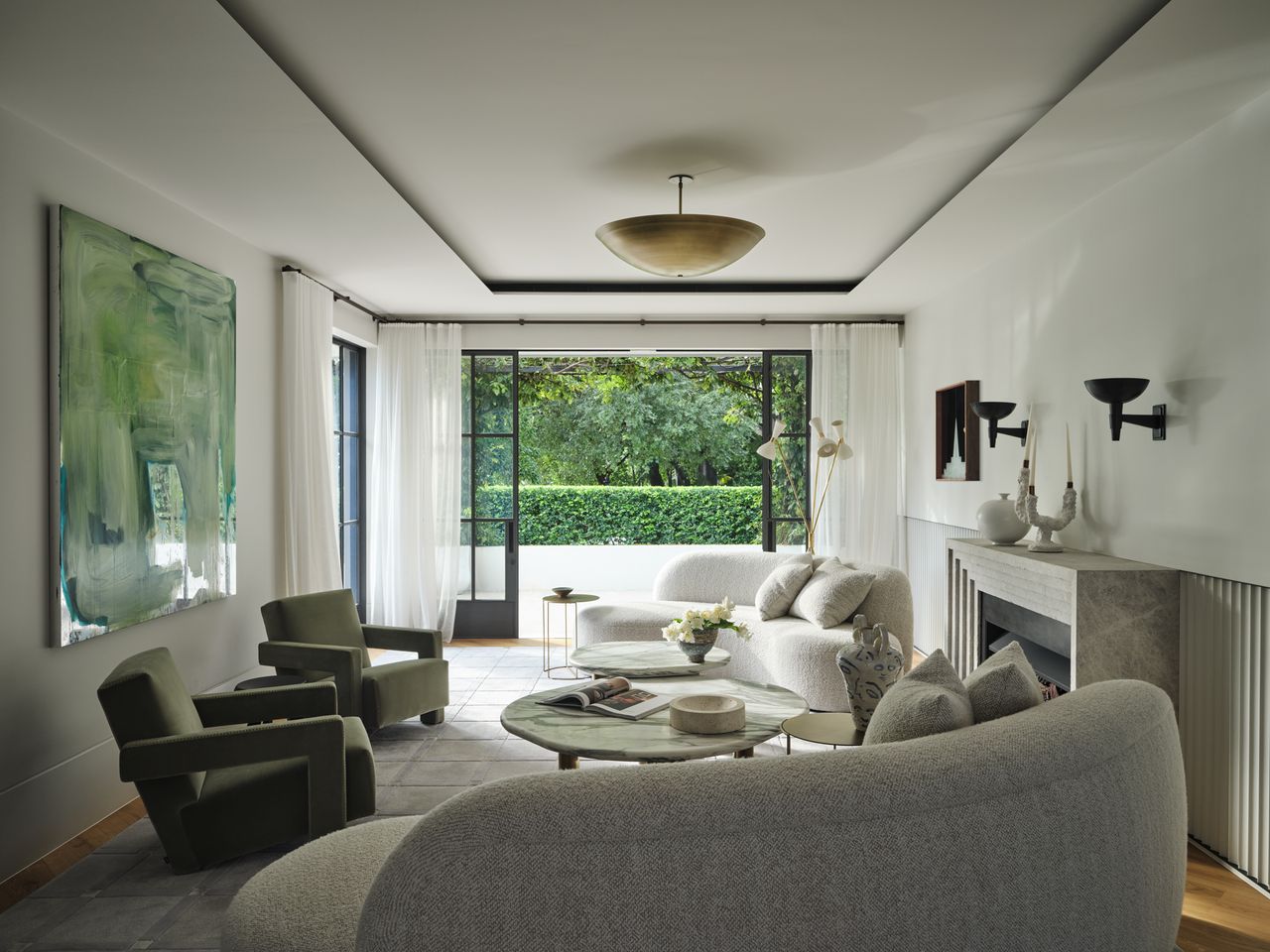 Monsieur G Living room with curved boucle sofas and sculptural olive-green armchairs around a round green onyx coffee table, framed by black-framed French doors opening to a garden, with a large abstract green painting, a stone fireplace, and a brass bowl-shaped ceiling pendant.