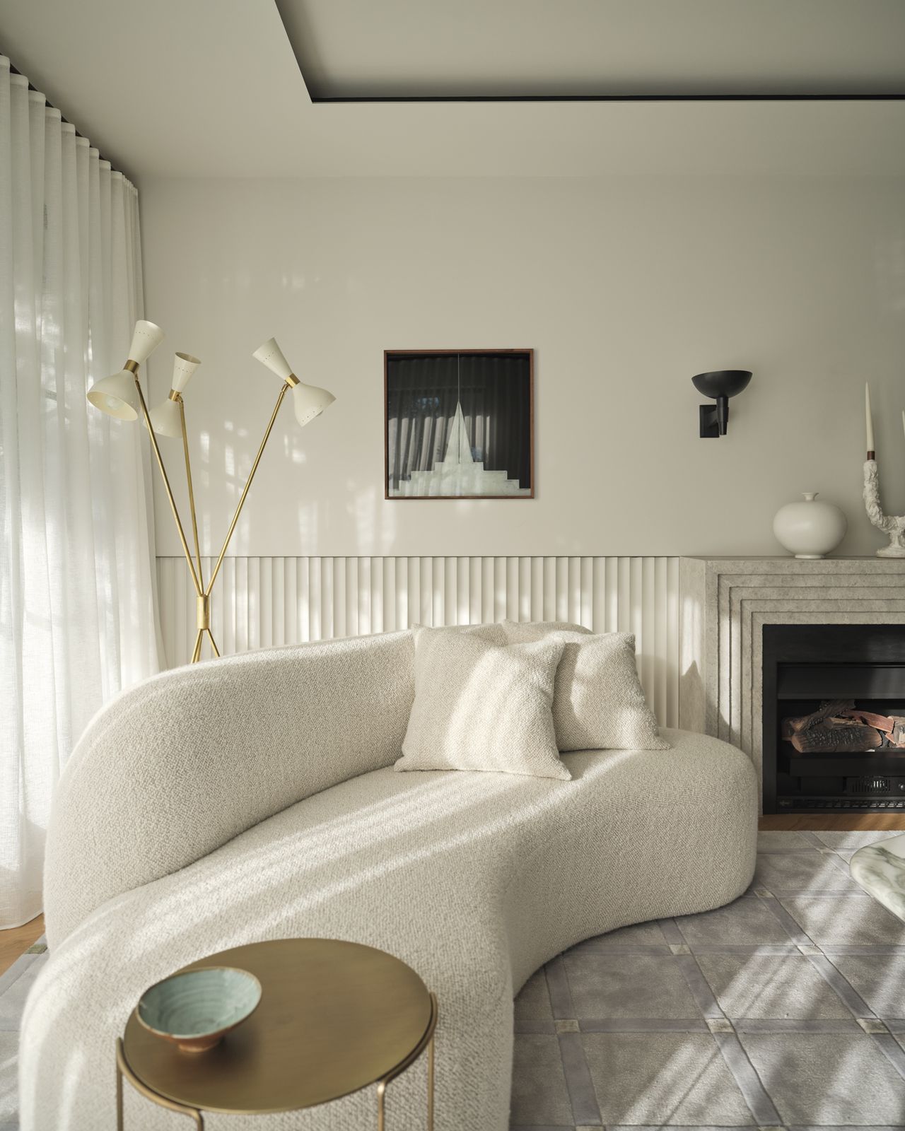Monsieur G Curved boucle sofa in a living room with fluted wall panelling, a stone fireplace, a brass sputnik-style floor lamp, a small framed artwork, and a brass side table topped with a ceramic bowl.