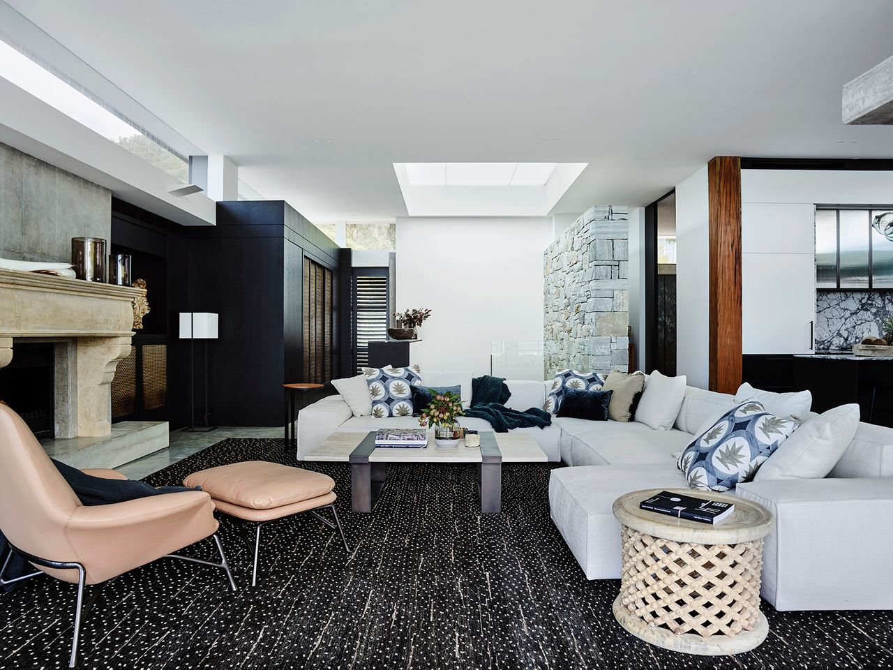 Killcare Living room with a large white sectional sofa and patterned cushions around a stone coffee table and a lattice-carved side table, an ornate stone fireplace, tan leather lounge chair and ottoman, and a natural stone feature column near the kitchen beyond