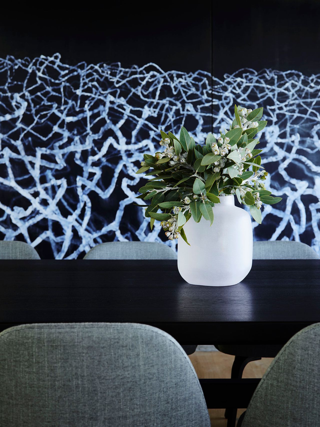 Killcare White ceramic vase with native greenery on a black timber dining table, set against a dark abstract artwork with a fine tangled linear pattern, framed by grey upholstered dining chairs