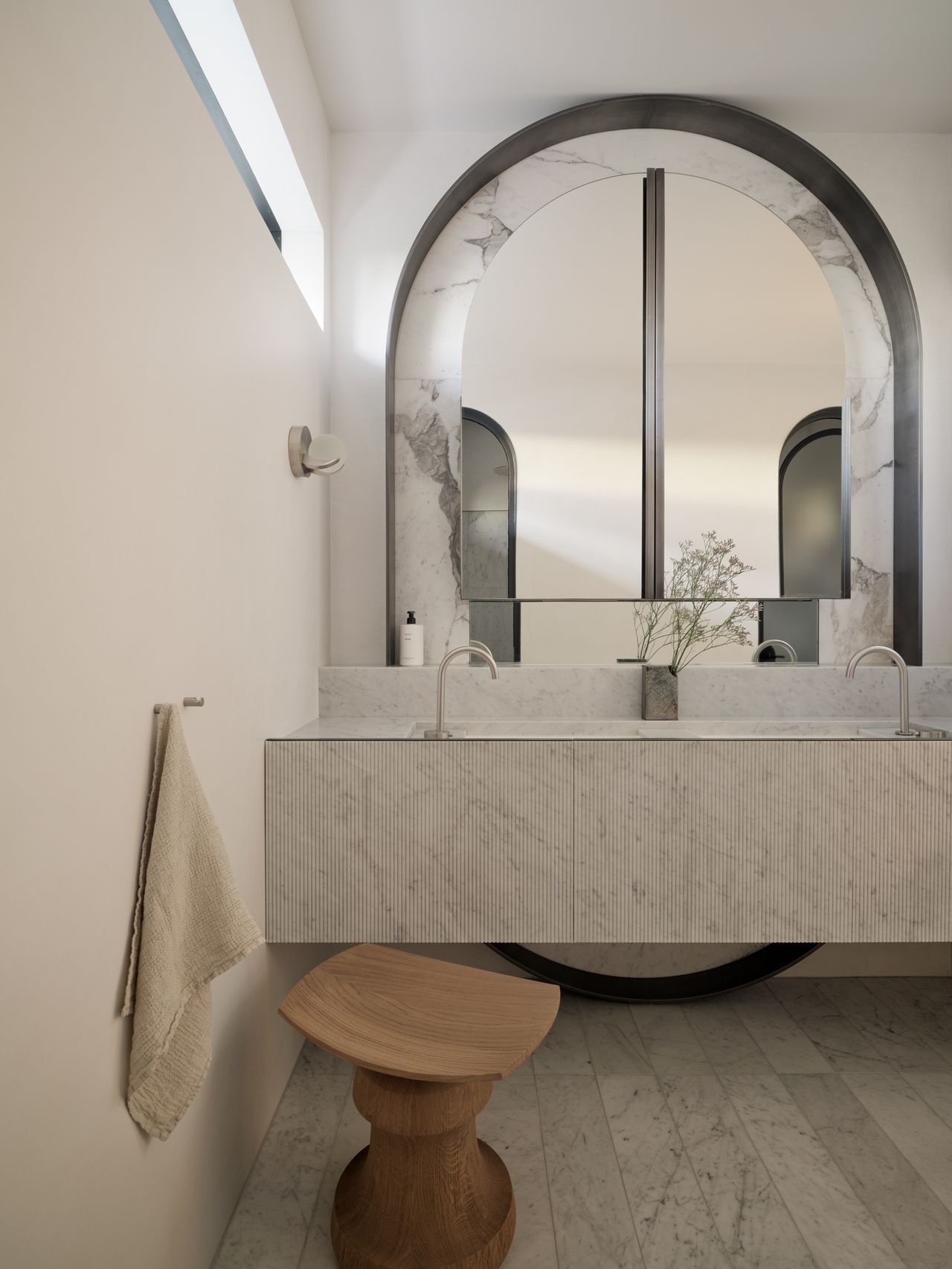 Monsieur G Bathroom with a fluted marble double vanity beneath a large arched mirror in a marble and blackened metal surround, styled with a small greenery sprig, and a sculptural timber stool on the marble-tiled floor