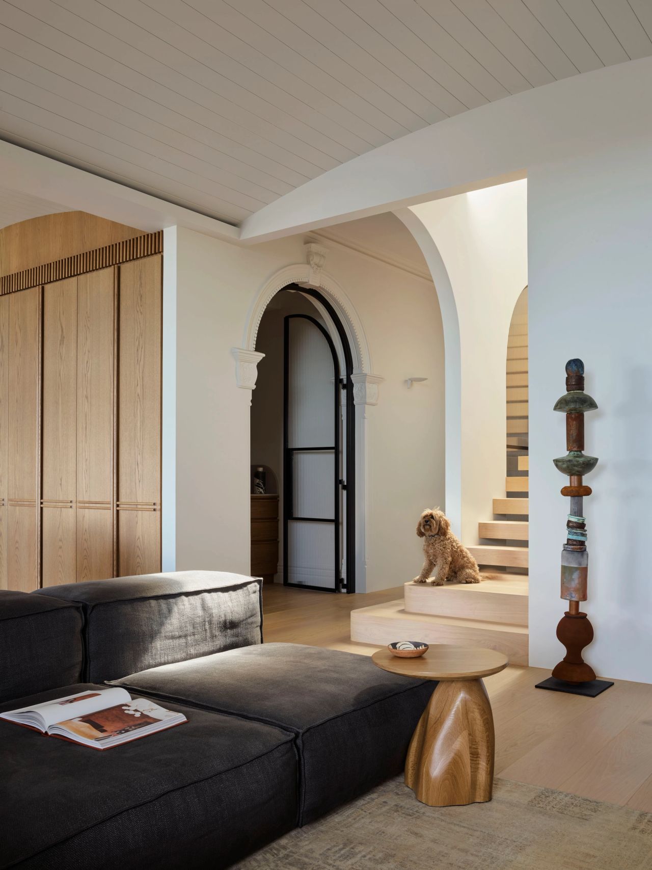 Ultramarine Living room with a charcoal linen sofa, sculptural wood side table, and a tall totemic ceramic sculpture, opening through an archway to a staircase where a curly-coated dog sits
