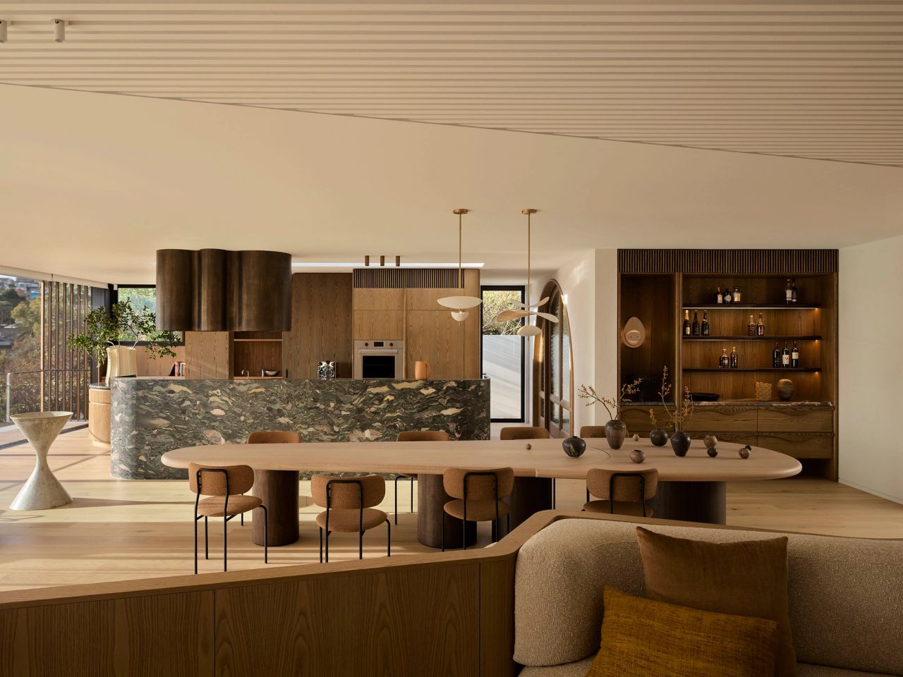 Patchwork Open-plan dining and kitchen space featuring a sculptural timber dining table with upholstered chairs, positioned in front of a stone kitchen island. Warm timber cabinetry, integrated appliances and a built-in display cabinet with decorative objects create a refined, natural palette. Floor-to-ceiling windows bring in abundant natural light, while soft furnishings and layered timber finishes create a warm, contemporary interior.