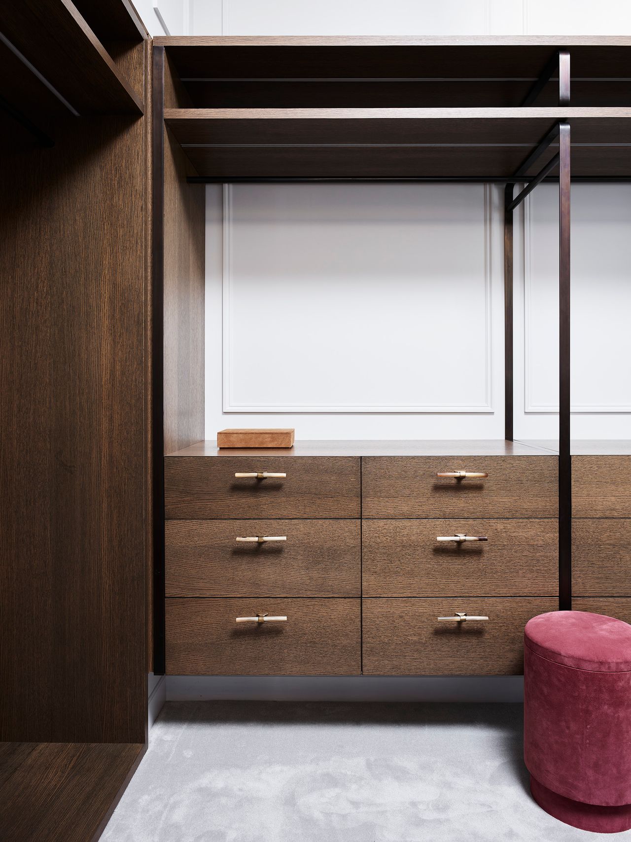 Bespoke walk-in wardrobe with dark timber cabinetry, brass drawer handles and a burgundy velvet ottoman.