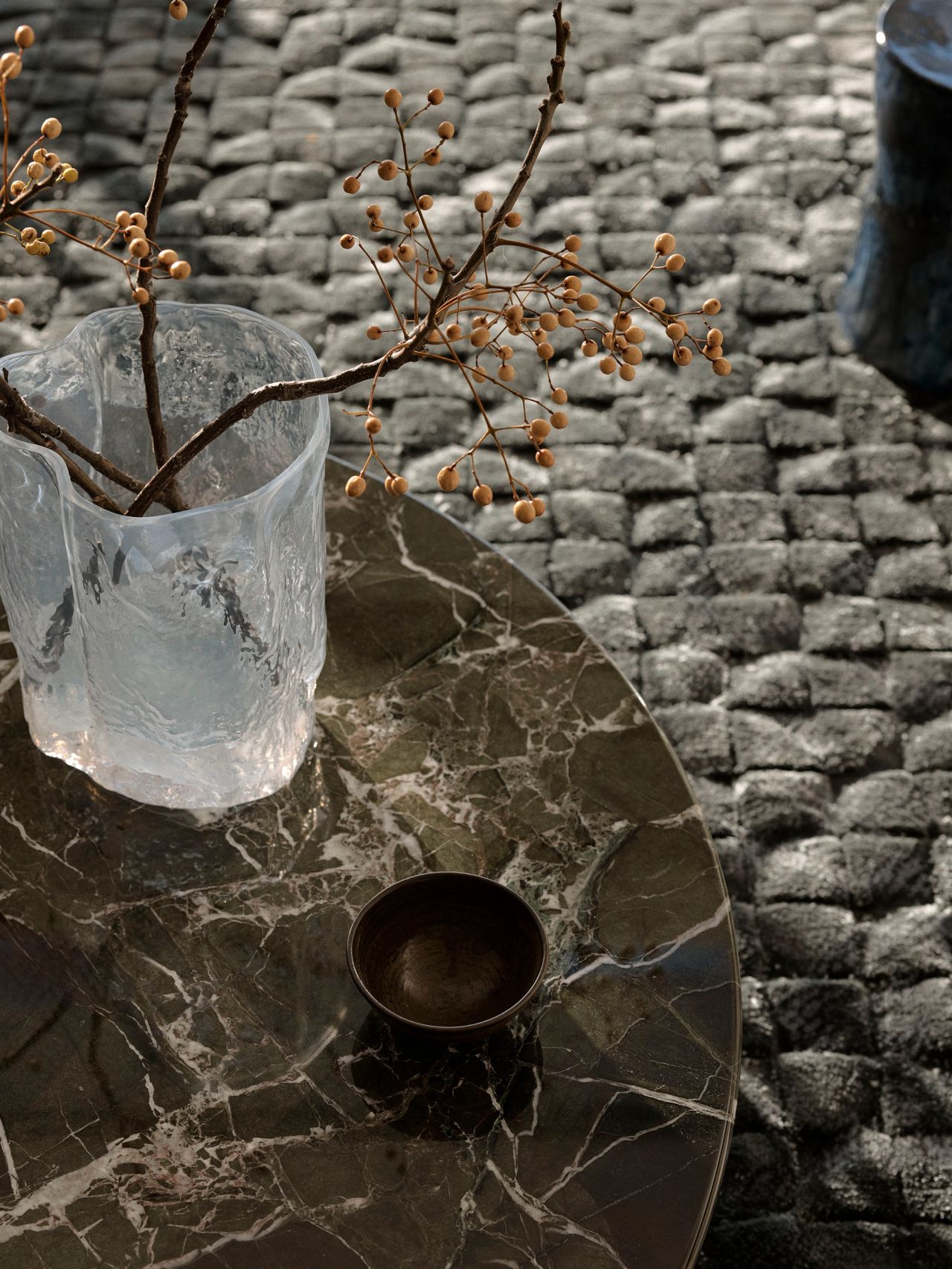 Sunset Textured glass vase with dried berry branches and a small bronze bowl on a dark veined marble table, set against a chunky woven charcoal rug