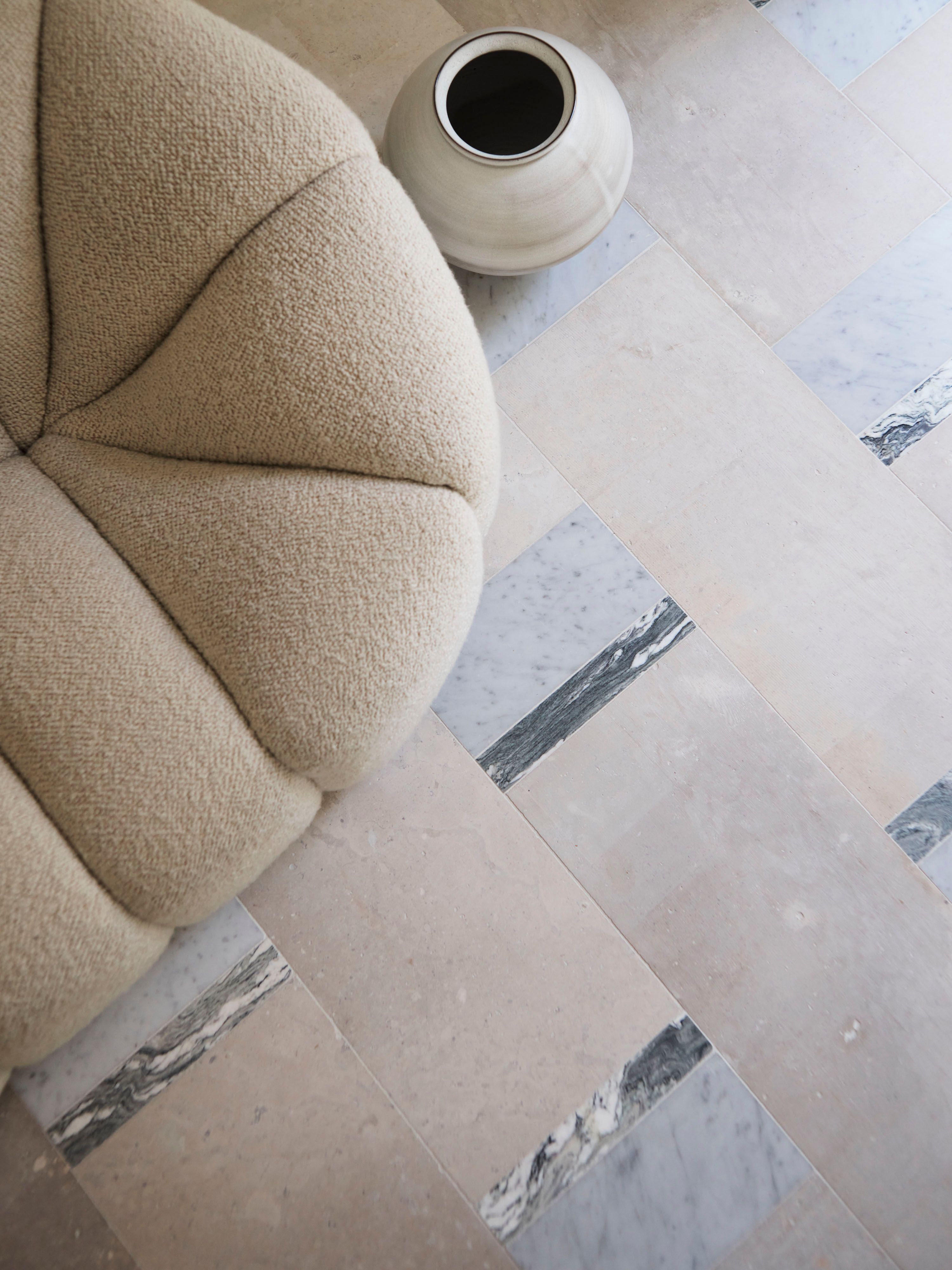 Ultramarine A close-up, top-down view of a beige boucle sofa's channel-tufted, petal-like cushion detail beside a round matte cream ceramic vase with a dark glazed opening. Both sit on a stone floor patterned with pale limestone tiles bordered by strips of grey-veined marble.