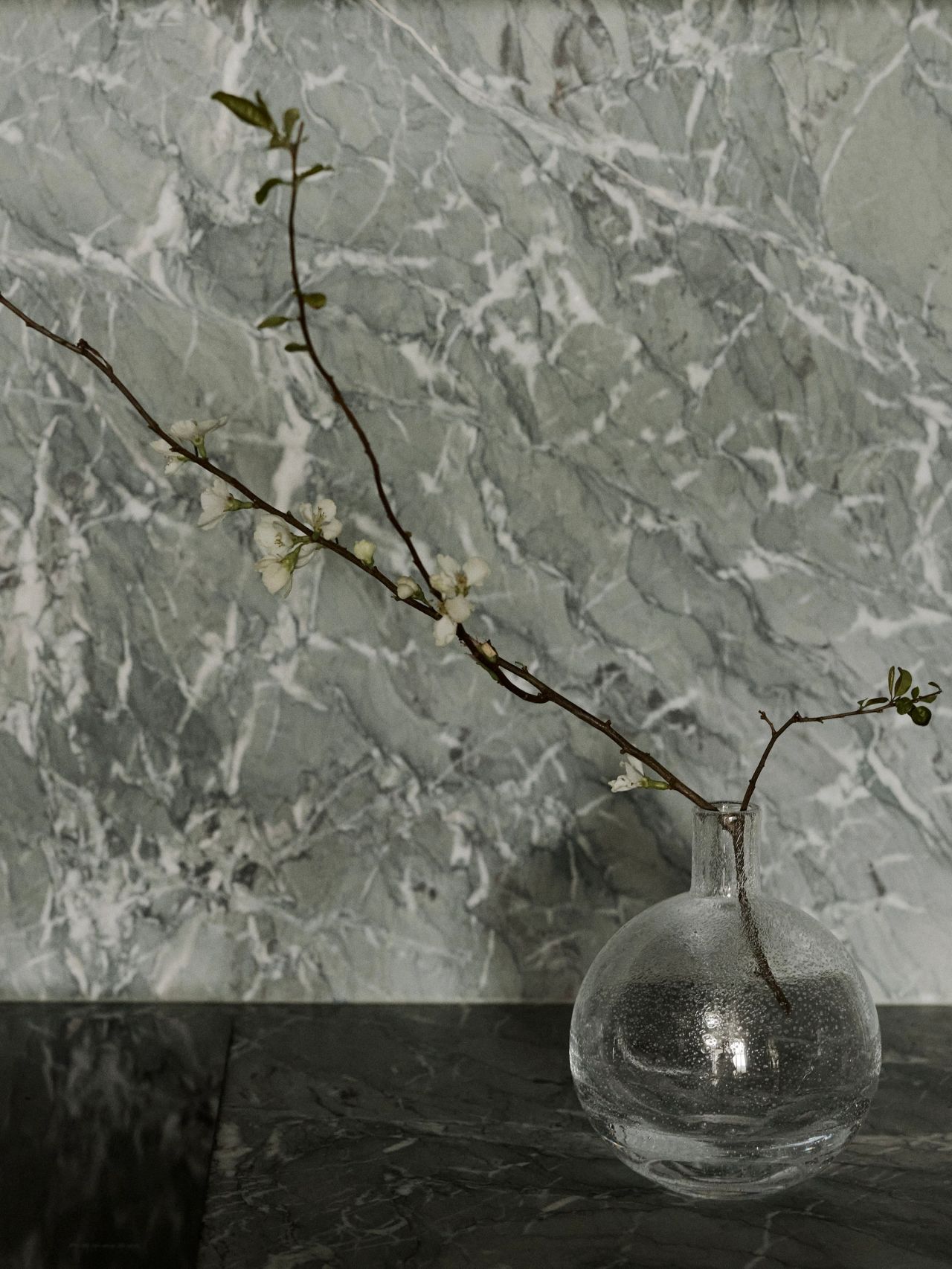 Sunset Round glass bud vase with a delicate flowering branch, set against a dramatic grey-green and white veined marble backdrop