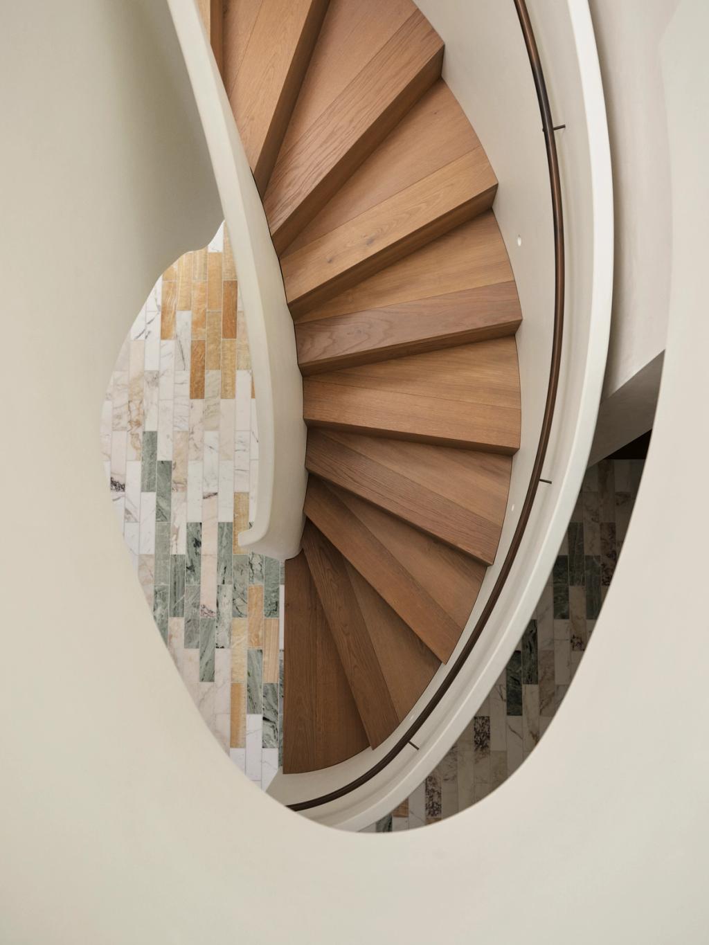 Dancing On The Ceiling Curved oval spiral staircase seen from above, framed by rendered walls and a mosaic tile floor in soft green, yellow, white and beige tones