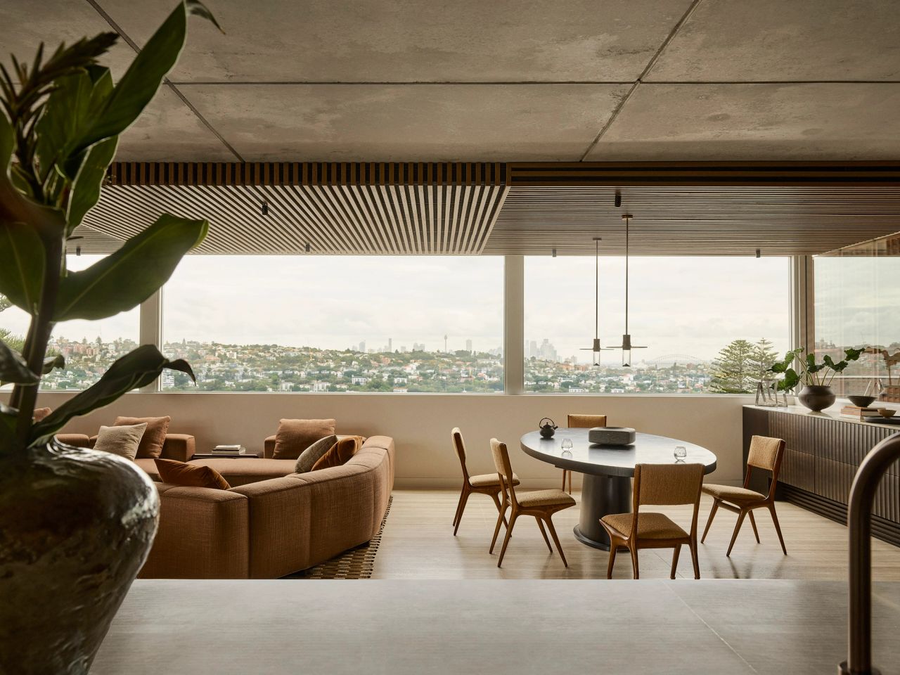 Orla Contemporary living room design open plan living and dining room blending Brazilian influences with a warm brown, black, and camel palette; black timber table with brown timber chairs. Timber batten ceiling detail. Views onto Sydney CBD and harbour