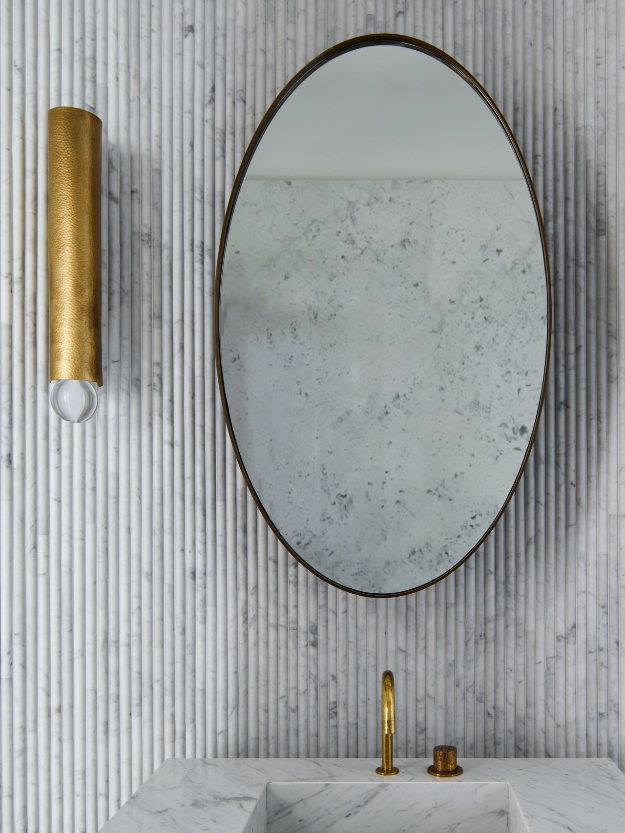 Detail of fluted white marble walls, oval mirror and warm brass fittings.