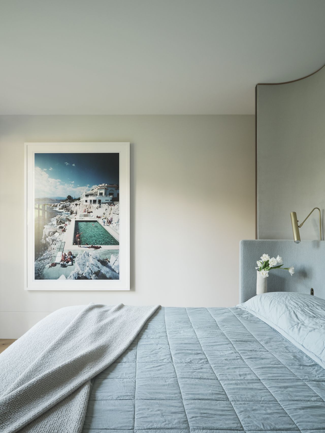 Monsieur G Primary Bedroom with a pale blue quilted bedspread, curved upholstered bedhead with brass reading light, styled with white flowers on the nightstand, and a large framed coastal photograph of a poolside villa on the wall.