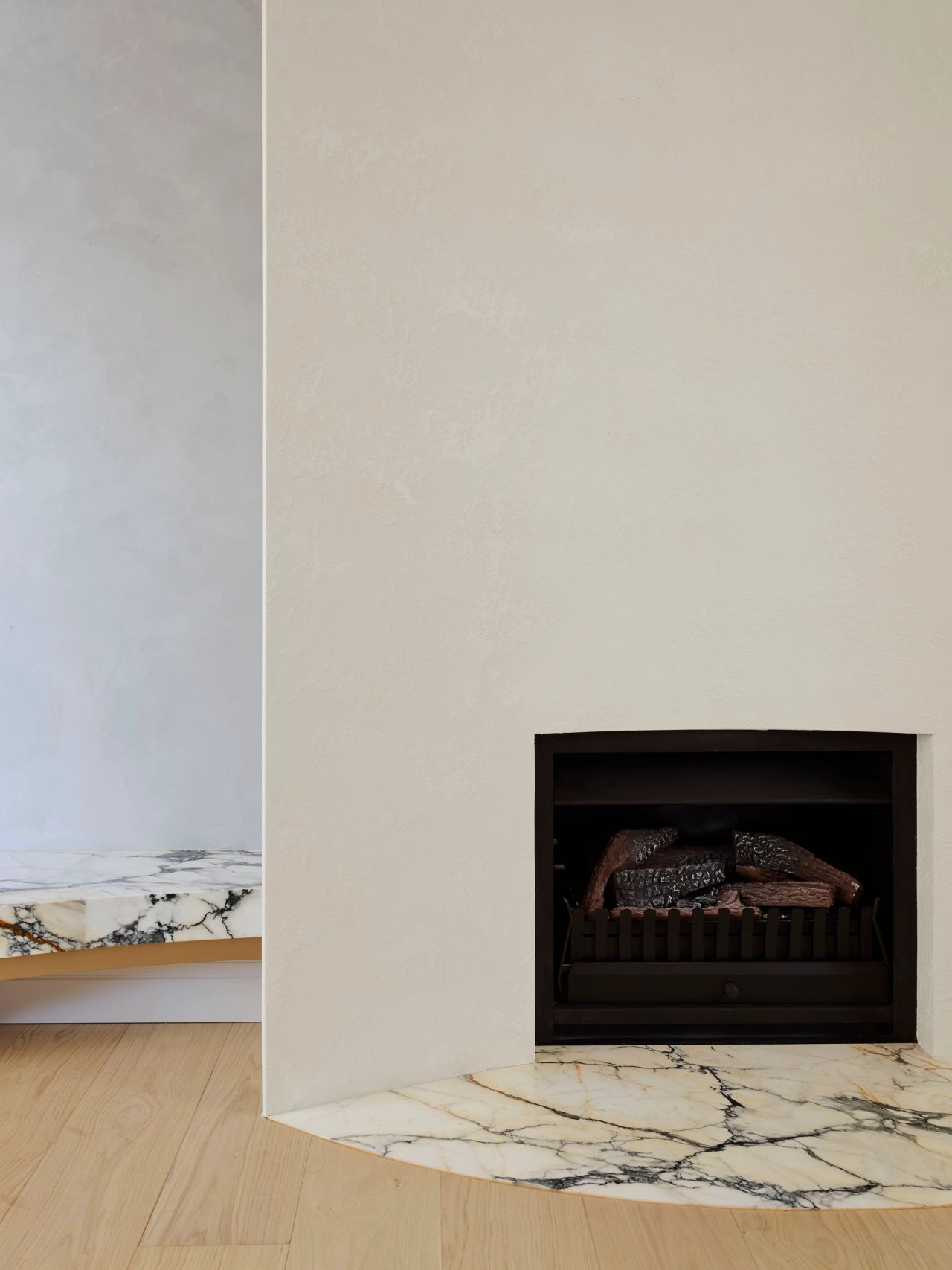 Ultramarine Curved plaster fireplace with a black inset grate holding logs, set on a dramatic veined marble hearth, with a matching marble bench visible in the background.