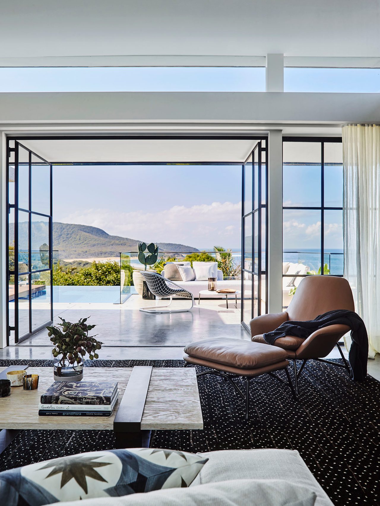Killcare Living room with black-framed glass doors opening onto a poolside terrace with coastal views, styled with a tan leather lounge chair and ottoman, a stone coffee table, and patterned cushions on a dark textured rug