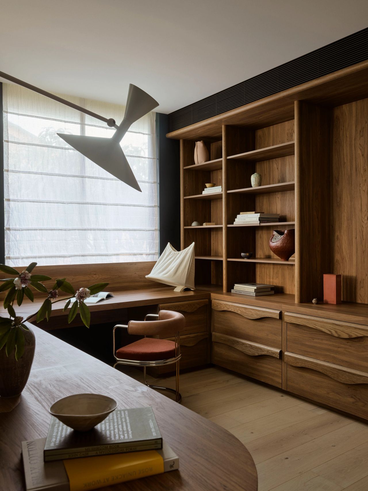 Patchwork Study nook with a built-in wooden desk beneath a sheer curtained window, a sculptural black arrow-shaped wall lamp, an upholstered stool, and open wood shelving styled with books and ceramics.