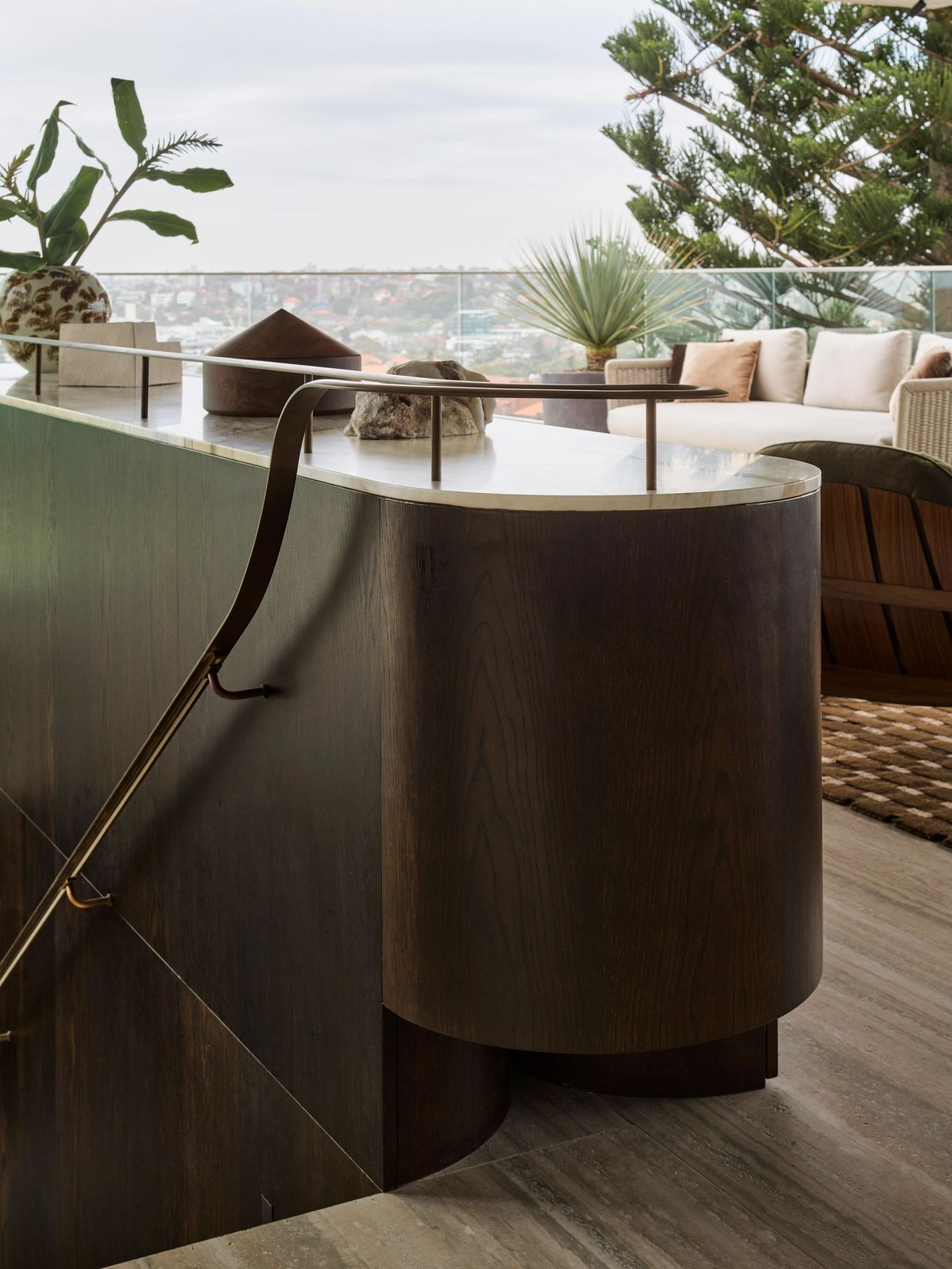 Orla contemporary staircase with Brazilian-influenced dark timber joinery featuring curved, fluted detailing, travertine flooring and a stone countertop