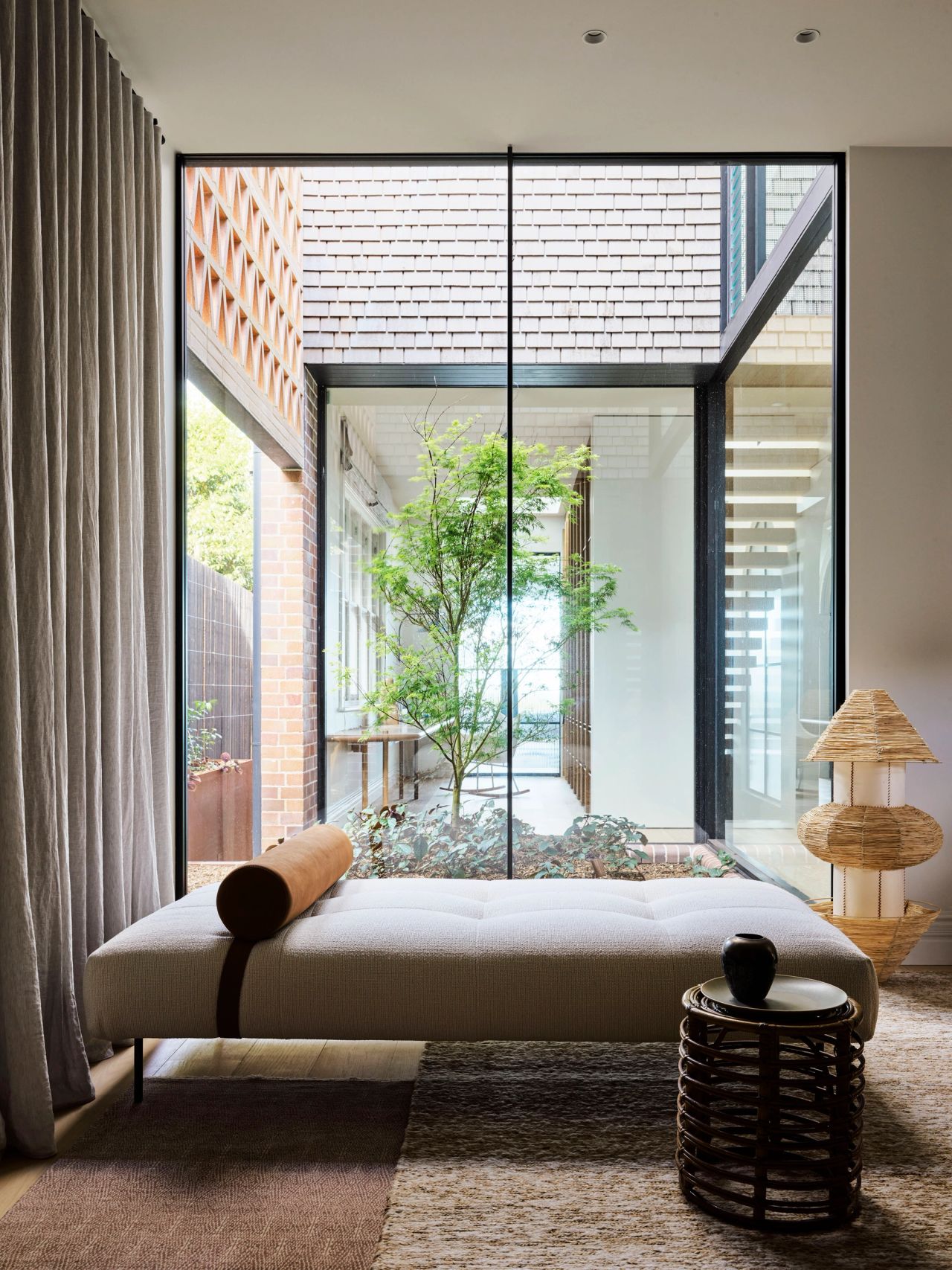 Ultramarine Upholstered daybed with a bolster cushion positioned beside floor-to-ceiling glass overlooking an internal courtyard tree, with a woven side table and sculptural lamp nearby