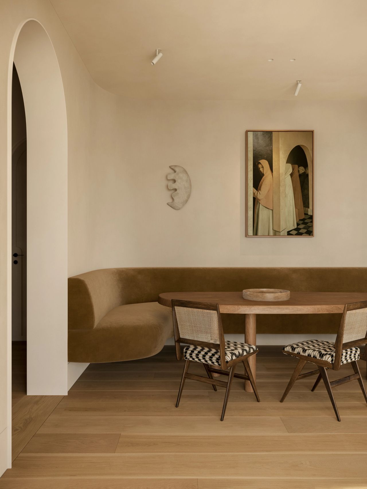 Lady M Breakfast nook with a curved olive-green velvet banquette wrapping an oval timber table, paired with cane-back chairs upholstered in black-and-white check. A figurative portrait painting and sculptural moon-shaped wall piece hang above, with an arched doorway to the left