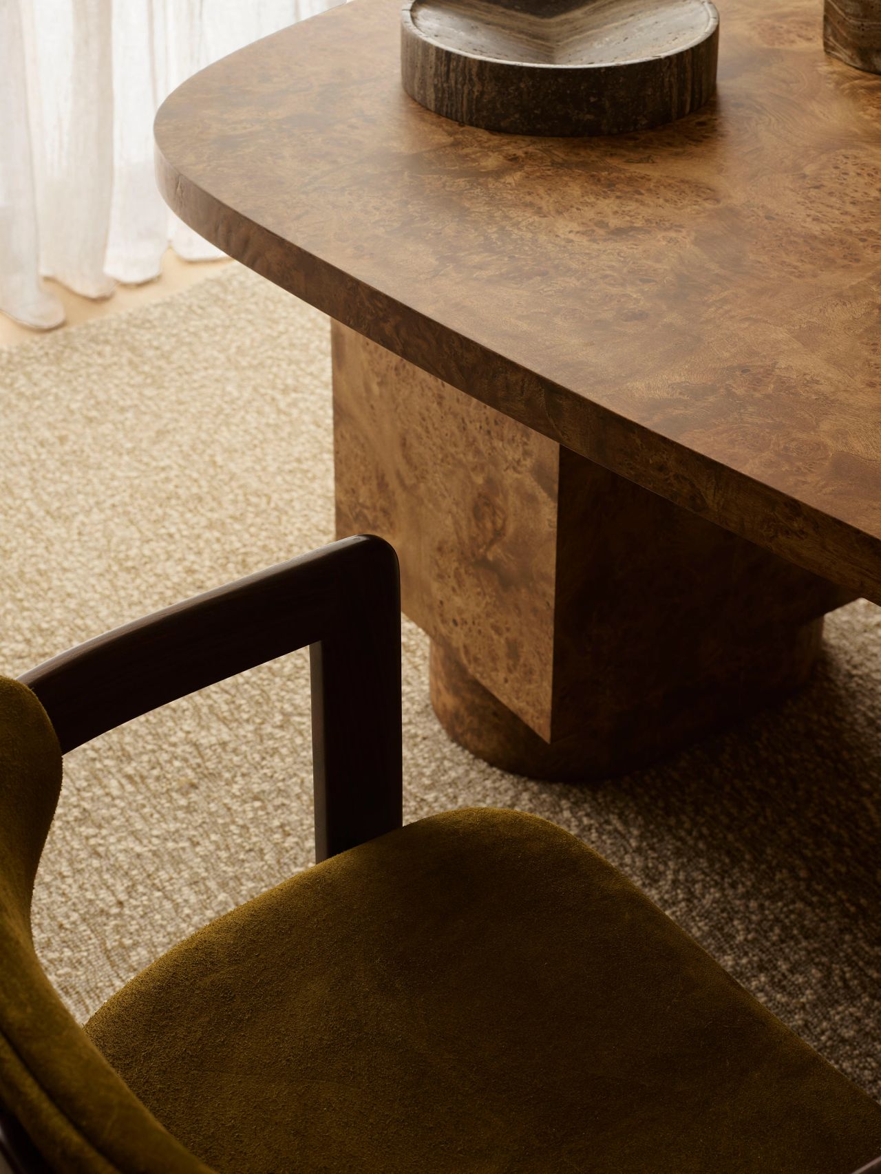 Patchwork Close-up of a burl wood desk with sculptural pedestal base, paired with an olive velvet armchair on a textured cream rug, with sheer curtains in the background.
