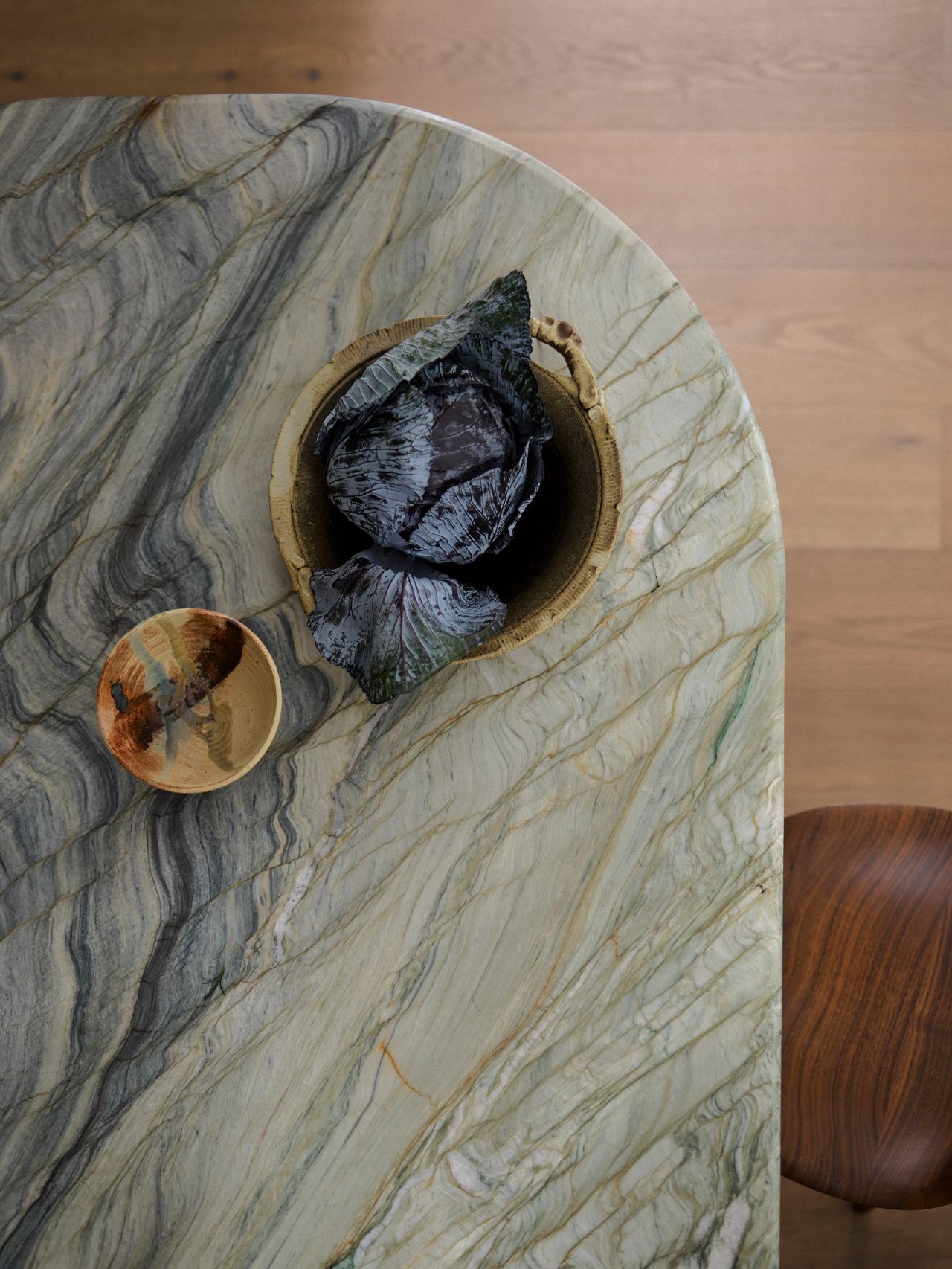 Dancing on the Ceiling Overhead view of a swirling green, blue, grey-veined marble table with a small sculptural ceramic object and a wooden bowl resting on the surface, beside a curved wooden stool