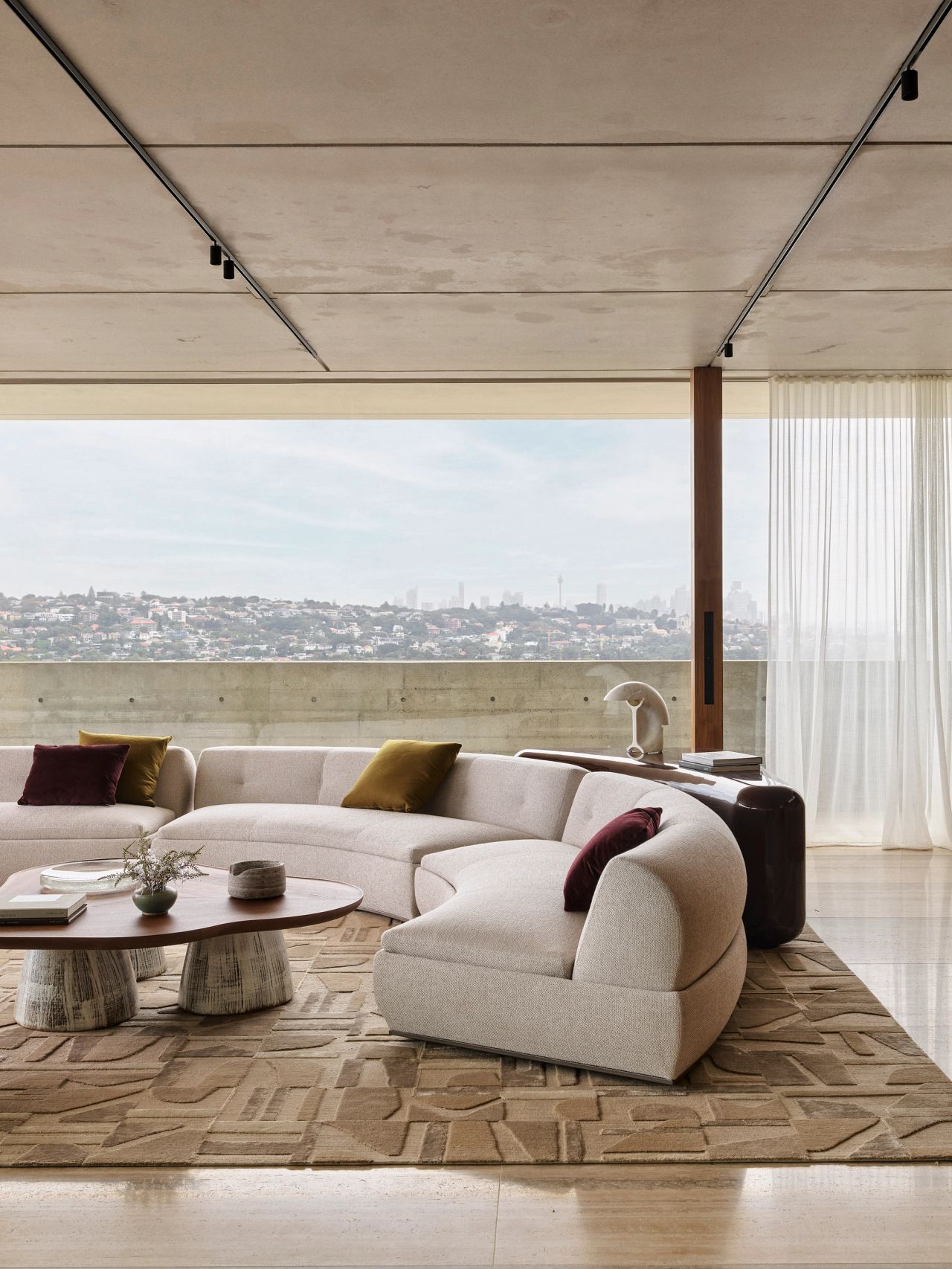 Orla contemporary open-plan living room blending Brazilian influences. A walnut coffee table with cream-glazed ceramic legs sits beside a Brazilian-style occasional chair with beige upholstery and timber arm detailing. A cream curved modular lounge sits on a geometric-patterned rug, with expansive views over Sydney's eastern suburbs and CBD, and walnut bar joinery featuring custom curved texture detailing.