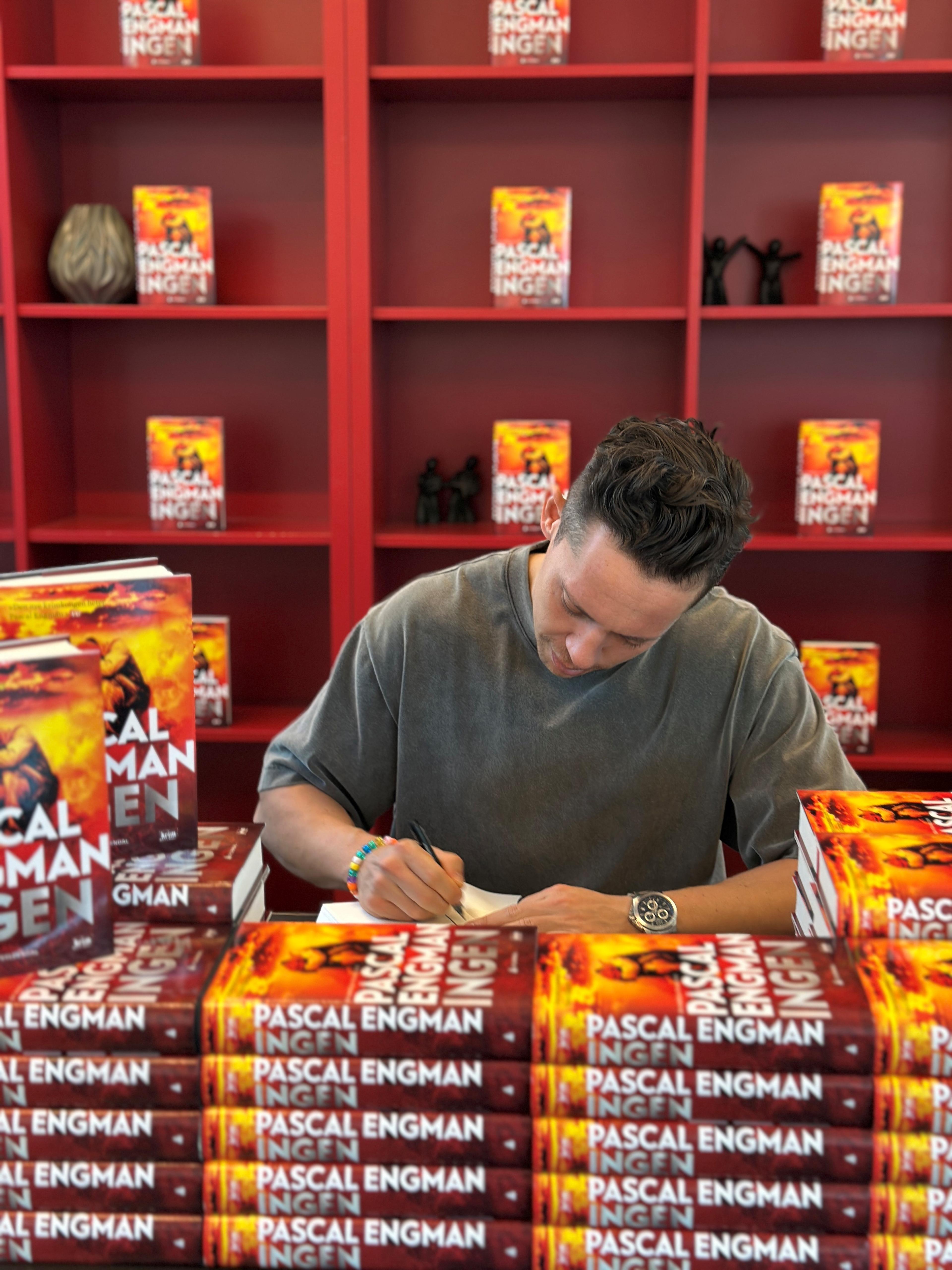 Pascal Engman sitter bak bokstabler med boka Ingen, og ser ned i en bok som han signerer