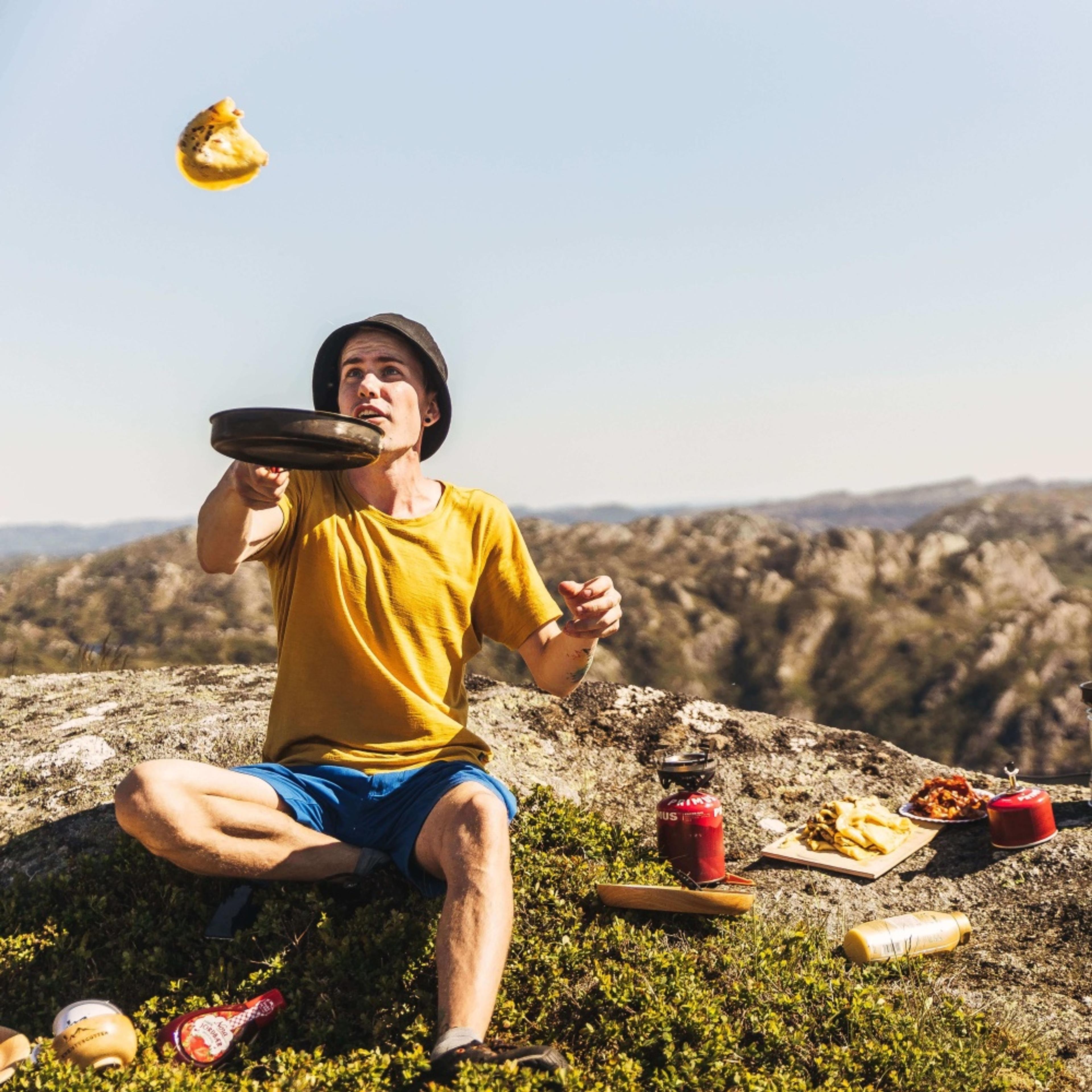 John Hobberstad sitter på en fjellknaus og kaster en pannekake fra en stekepanne opp i luften.