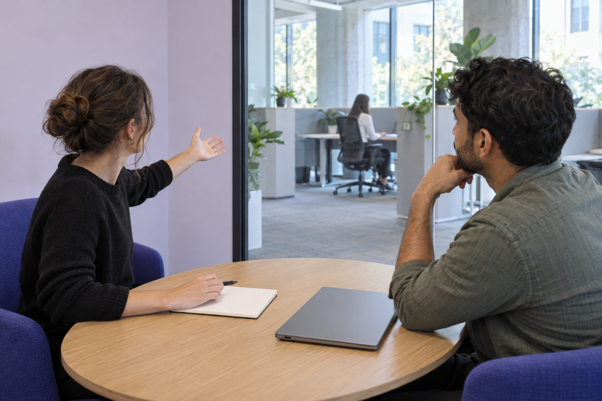 Two workplace coordinators discuss the nearby focus area from inside an enclosed meeting room.