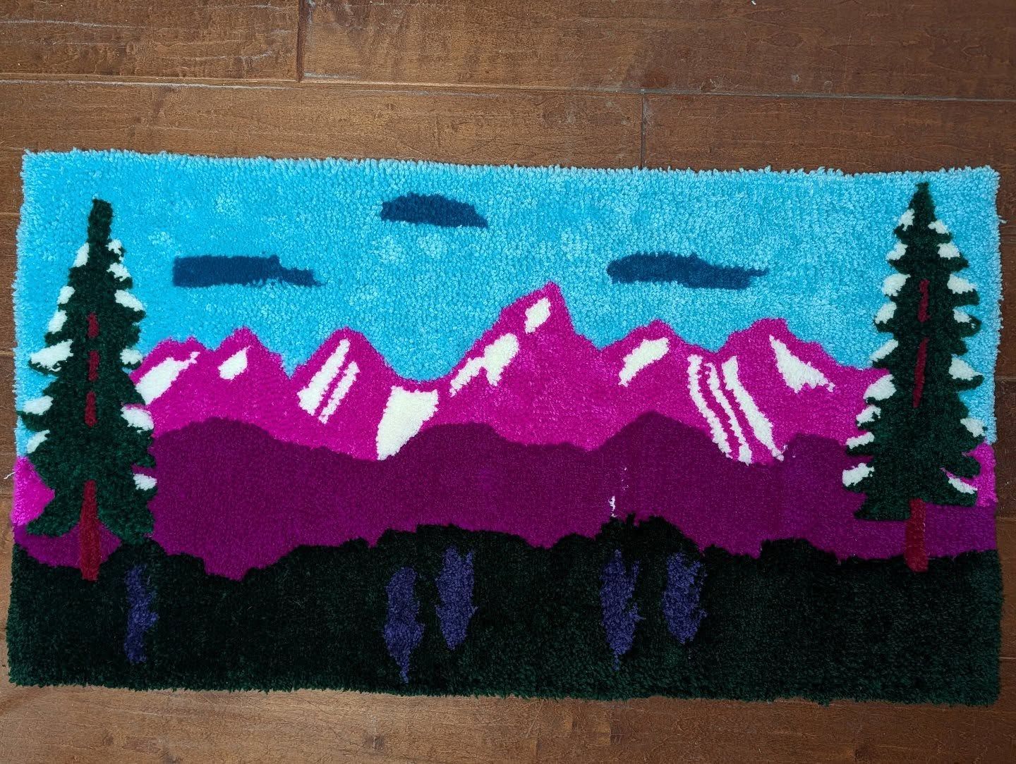 Rectangular hand-tufted rug depicting a vibrant mountain range in magenta and purple with pine trees, a teal sky, and dark clouds