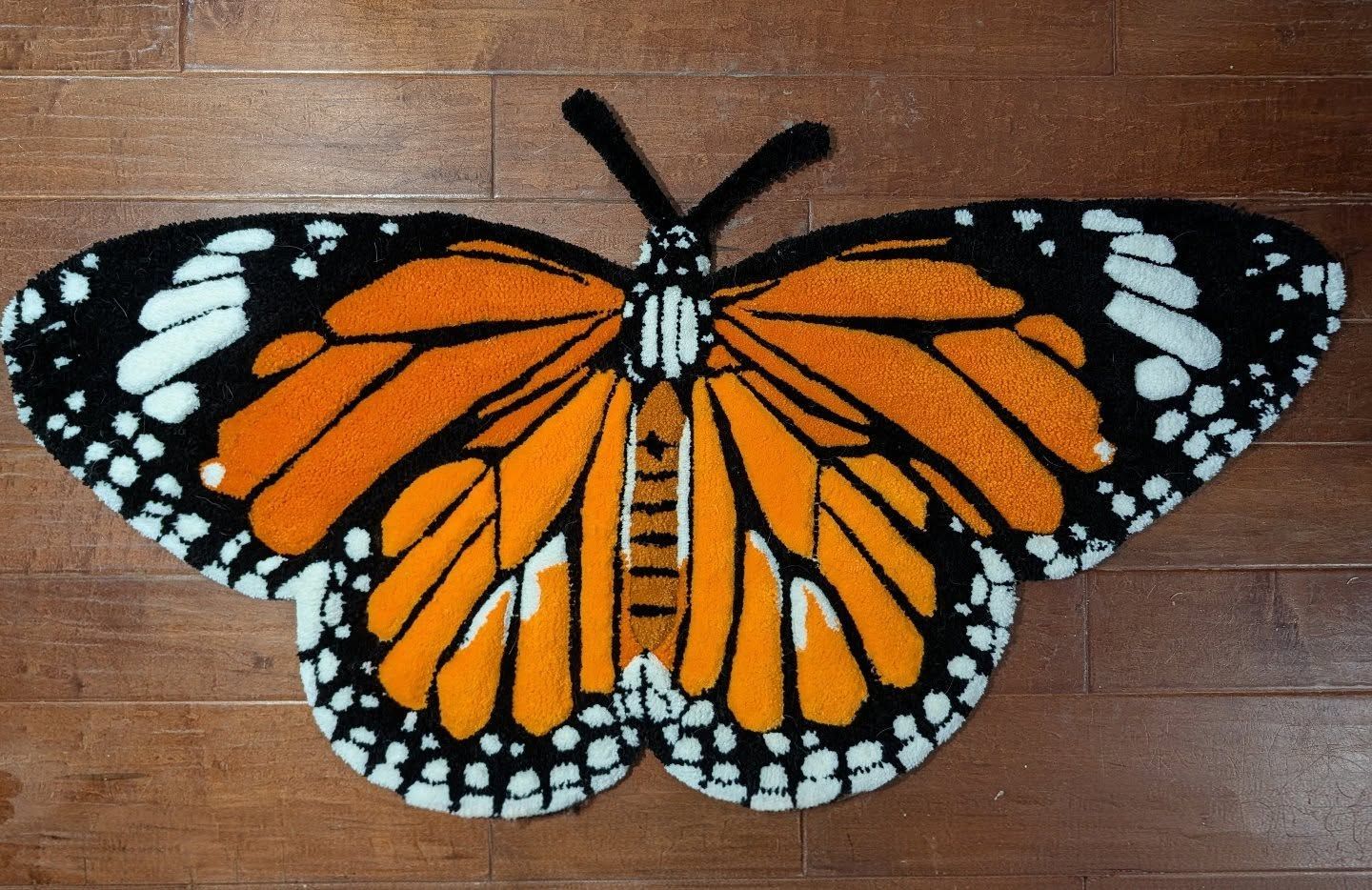 Hand-tufted rug shaped like a monarch butterfly with orange, black, and white wings on a dark wood floor