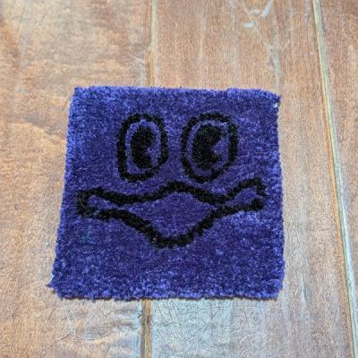 Small square hand-tufted rug in deep purple with a quirky black cartoon face with wide eyes and a squiggly mouth