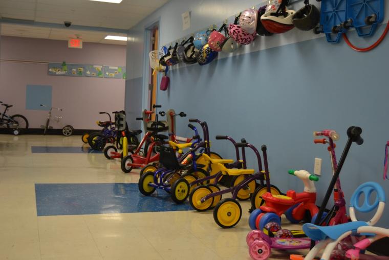 A corridor lined with adaptive tricycles and bicycles, with children's helmets hanging on a rail along the wall above them.
