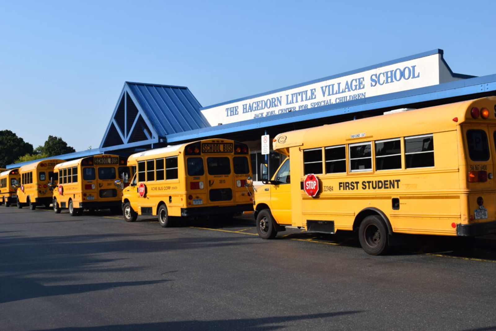 School buses lined up outside the school's main entrance, beneath the blue peaked roof and the sign reading The Hagedorn Little Village School, Jack Joel Center for Special Children.