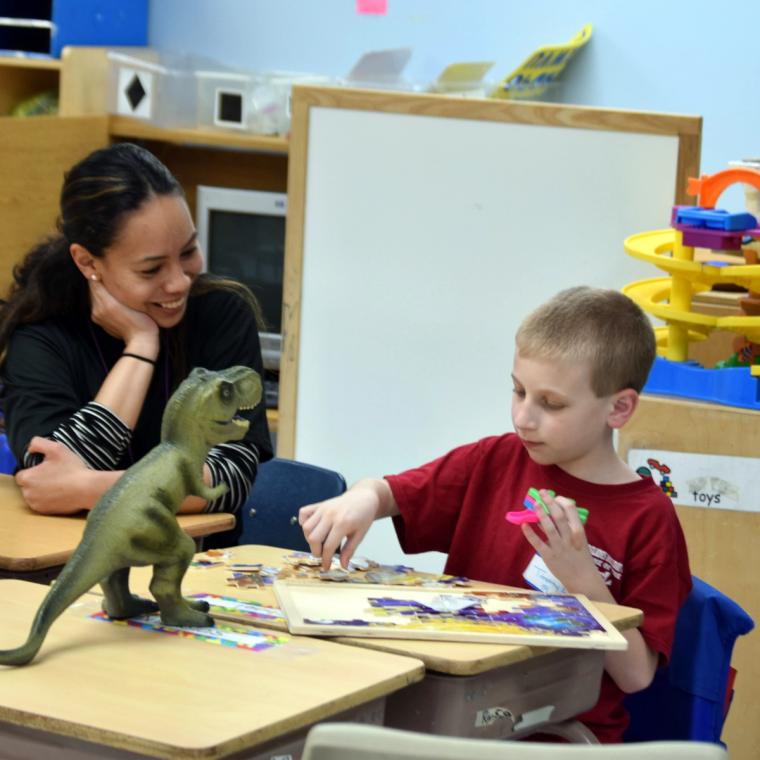 A teacher sits with a boy at a classroom table as he works on a jigsaw puzzle.
