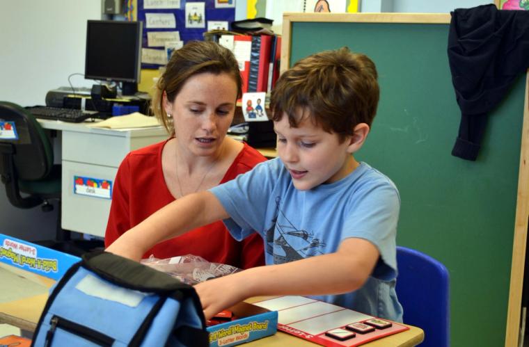 An evaluator sits beside a boy at a classroom table as he reaches into a bag next to a magnetic letter board.