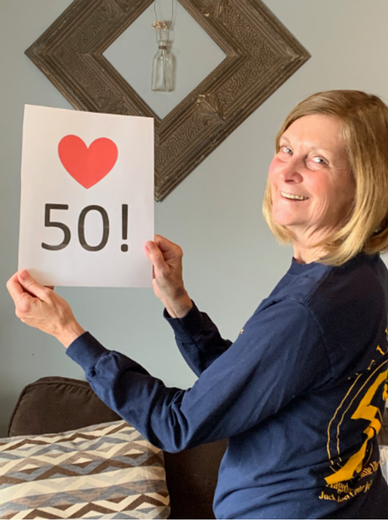 A woman in a Hagedorn Little Village School shirt smiling and holding up a sign with a red heart and “50!”, marking the school’s 50th anniversary
