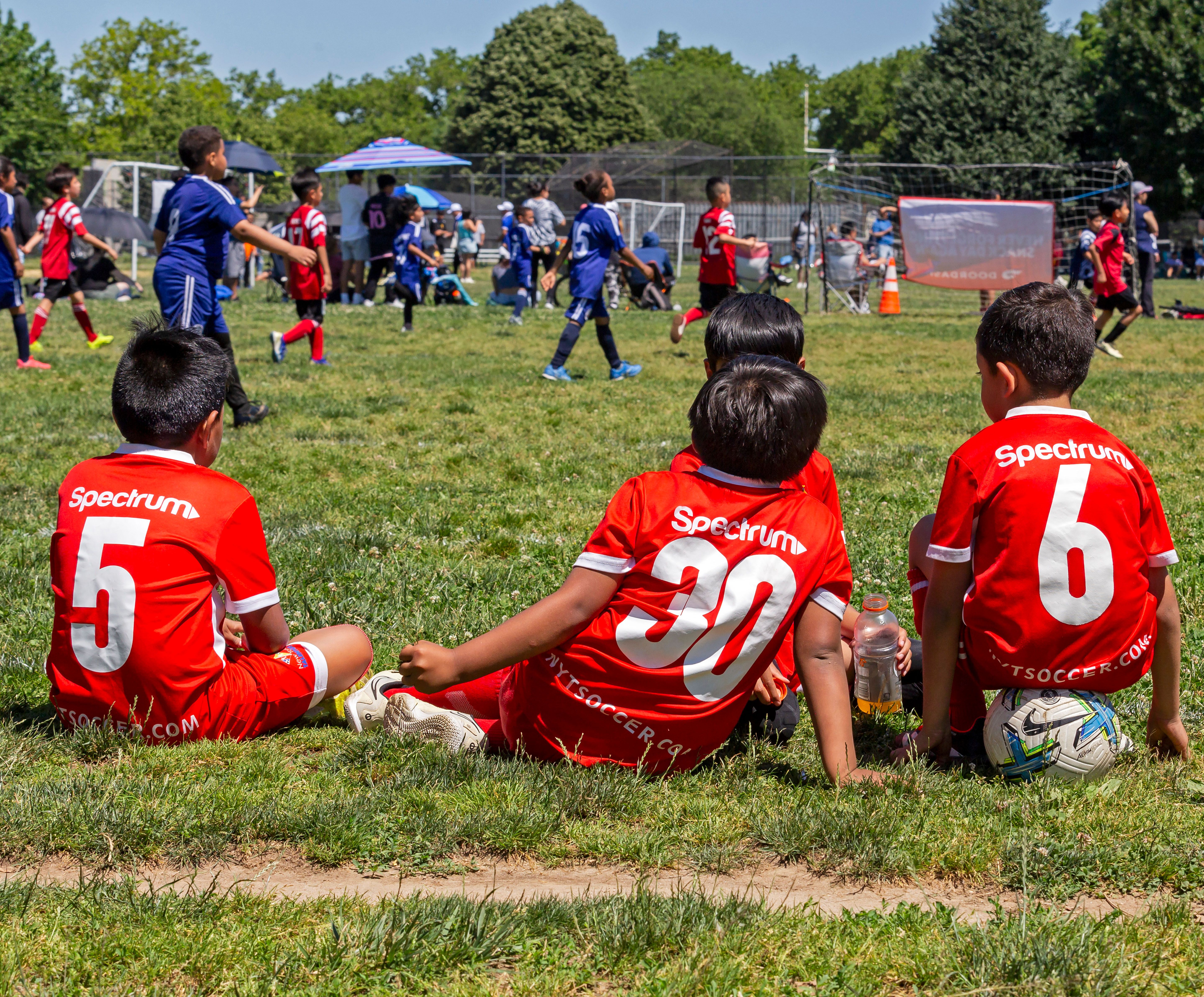Four young athletes sitting on the sidelines with their backs to camera; wearing red, Spectrum jerseys