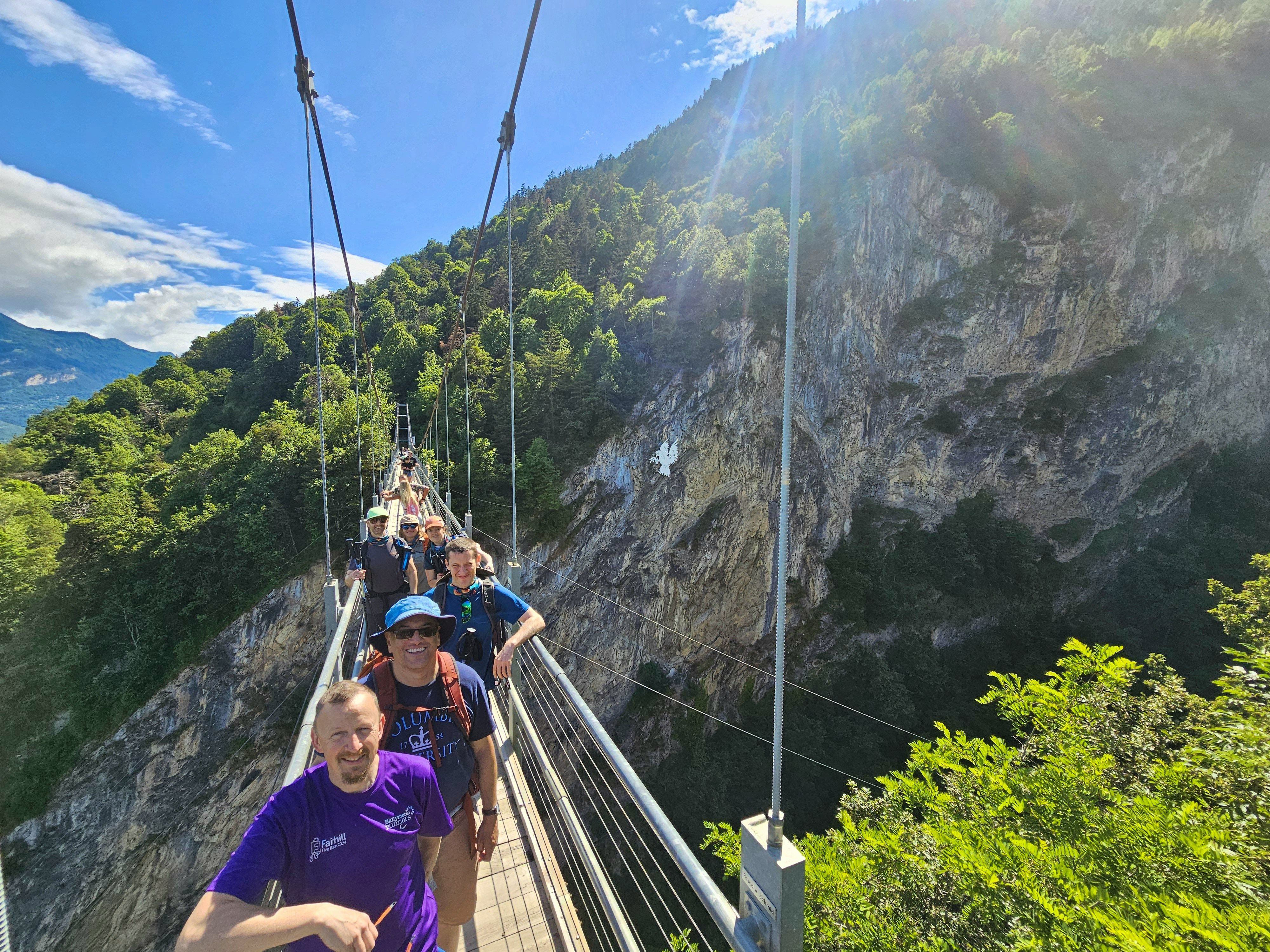 Group of smiling hikers on a suspension bridge with cliff face and valley behind