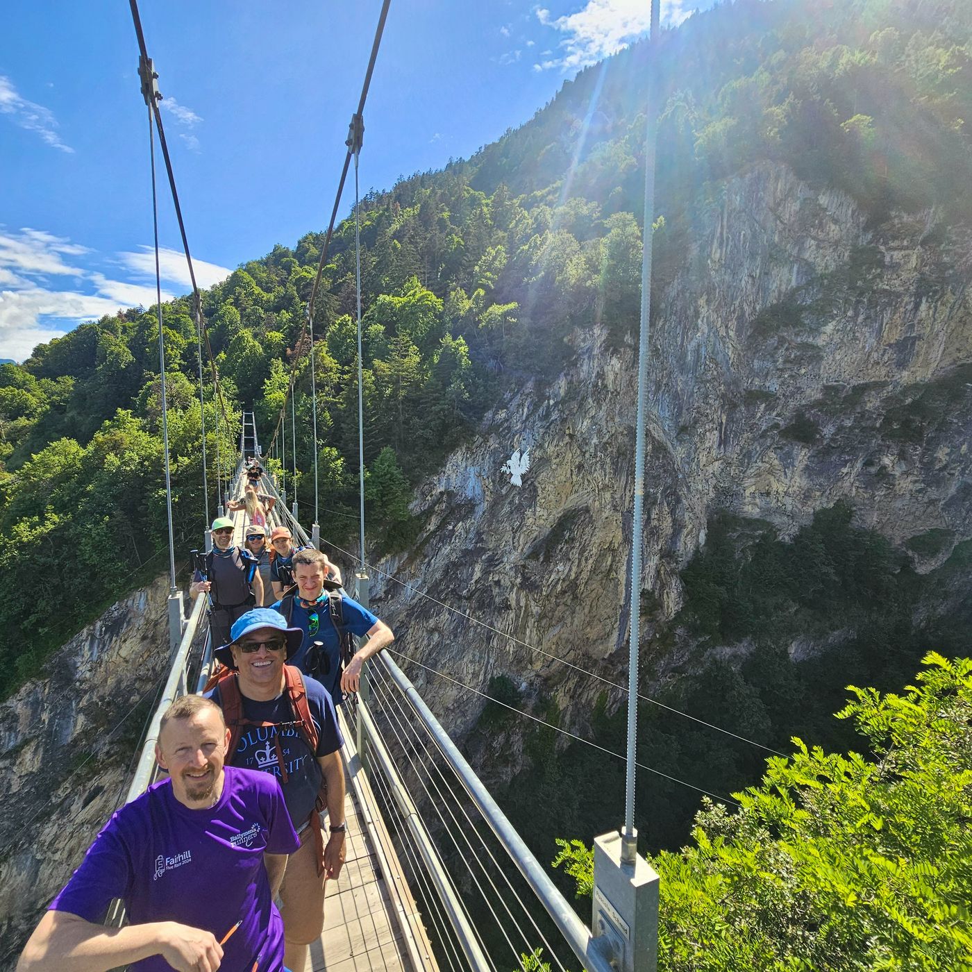 Group of smiling hikers on a suspension bridge with cliff face and valley behind