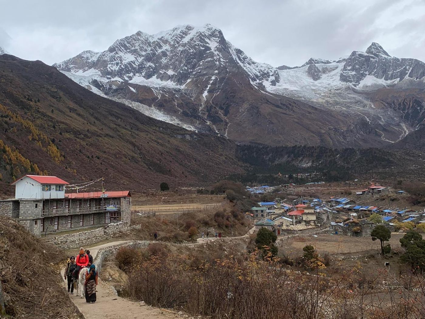 Trekkers approaching a mountain village beneath snowy Himalayan peaks