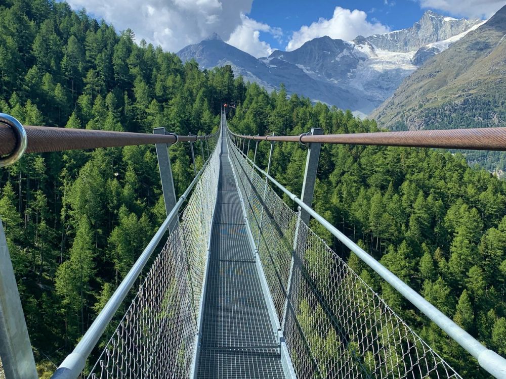 Charles Kuonen suspension bridge stretching into the distance above pine forest with snow-capped peaks