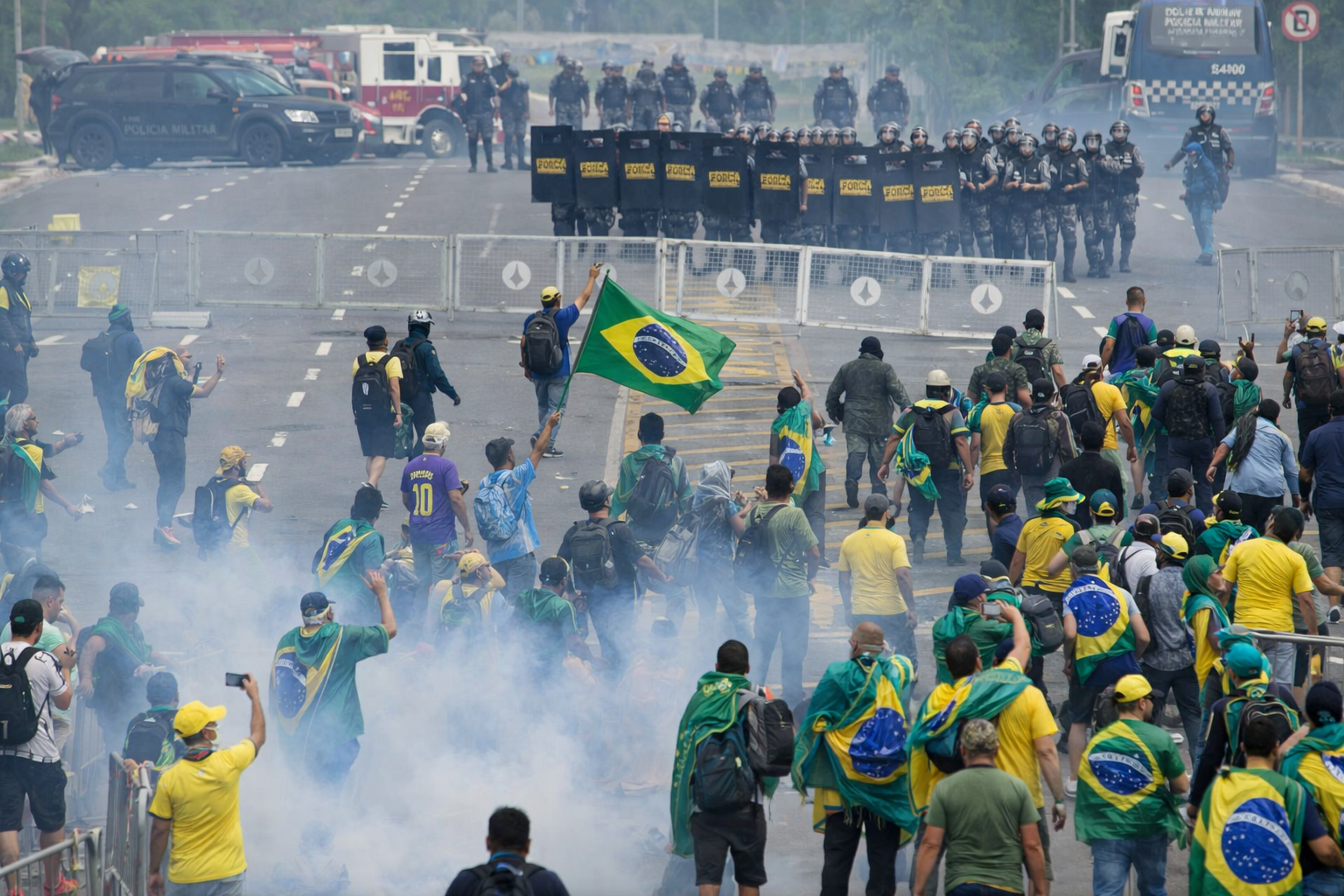 Crowd in yellow and green waving Brazilian flags faces a line of riot police with shields as tear gas fills the street in Brazil.
